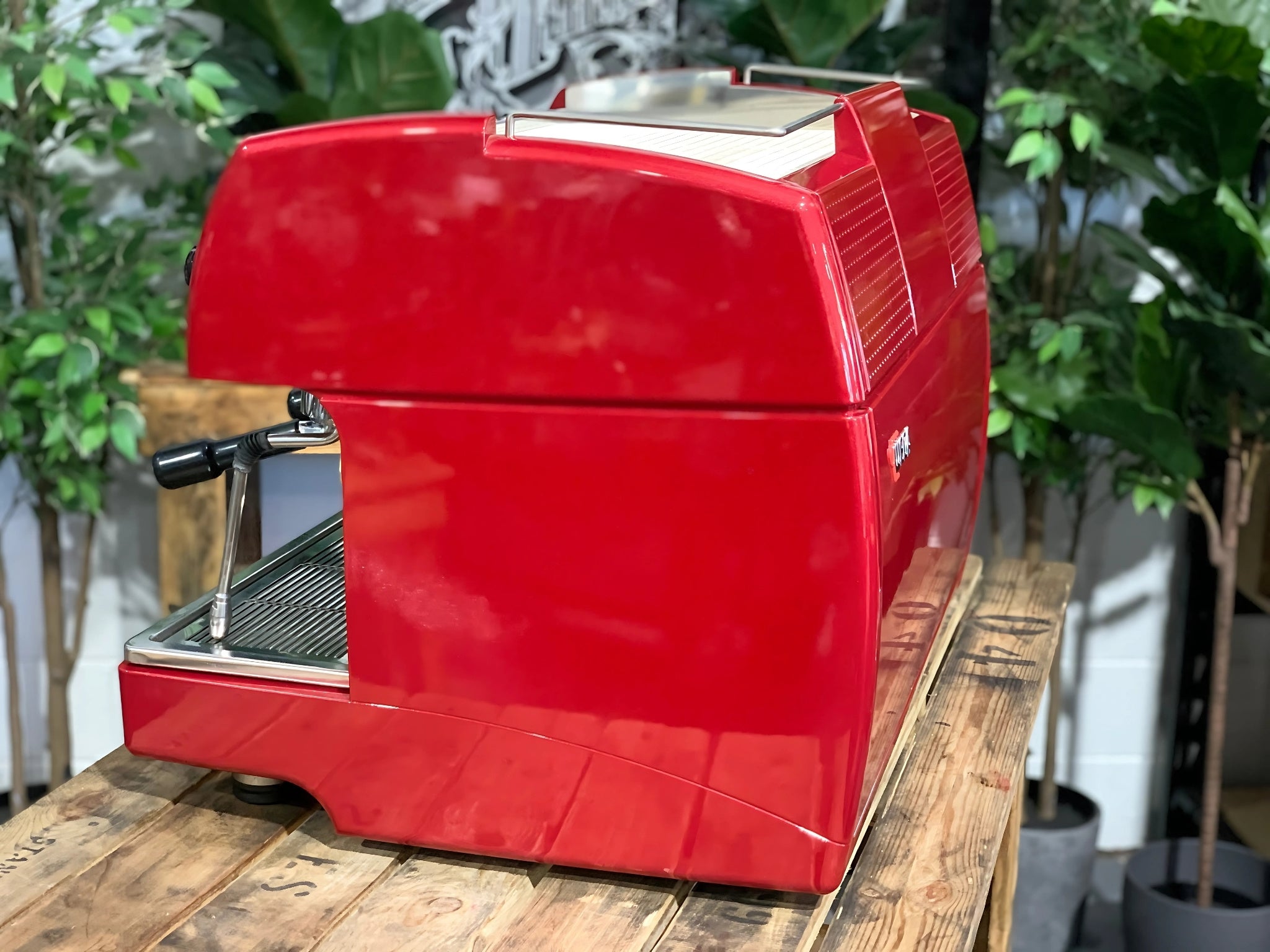 A Wega Sphera 2 Group Espresso Coffee Machine with Red finish sitting on a wooden bench. There is greenery in the background. The image is taken from the side of the machine.