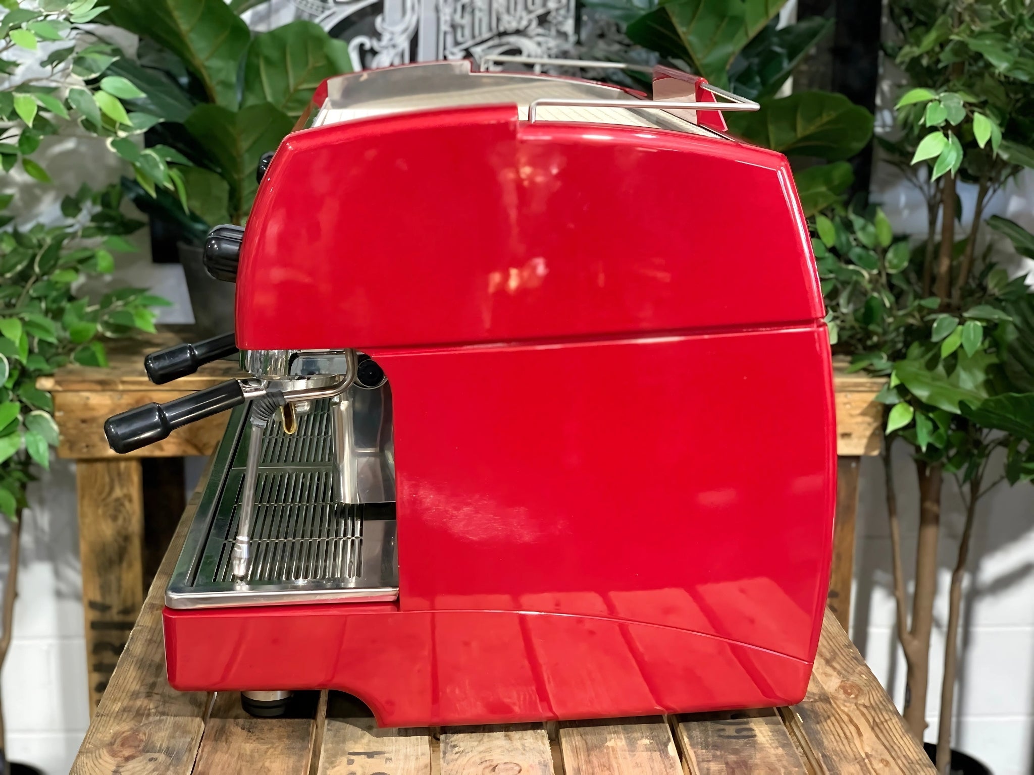 A Wega Sphera 2 Group Espresso Coffee Machine with Red finish sitting on a wooden bench. There is greenery in the background. The image is taken from the side of the machine.