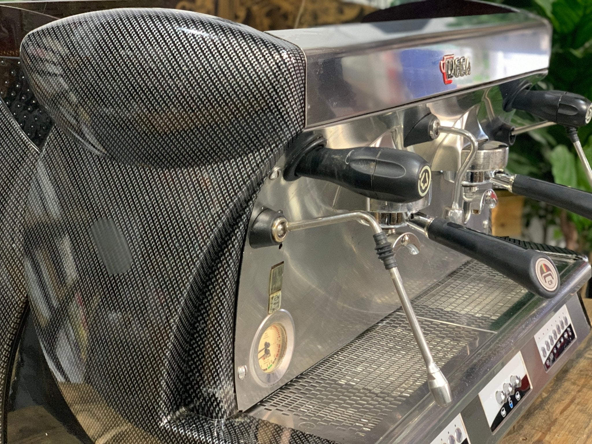 A Wega Vela 2 Group Espresso Coffee Machine with Carbon Fibre panels is sitting on a wooden bench. There is greenery in the background. The image is a close up image taken from the front of the machine and the machine is sitting on an angle so that you can see the front of the machine, the controls including buttons, group handles and logo.