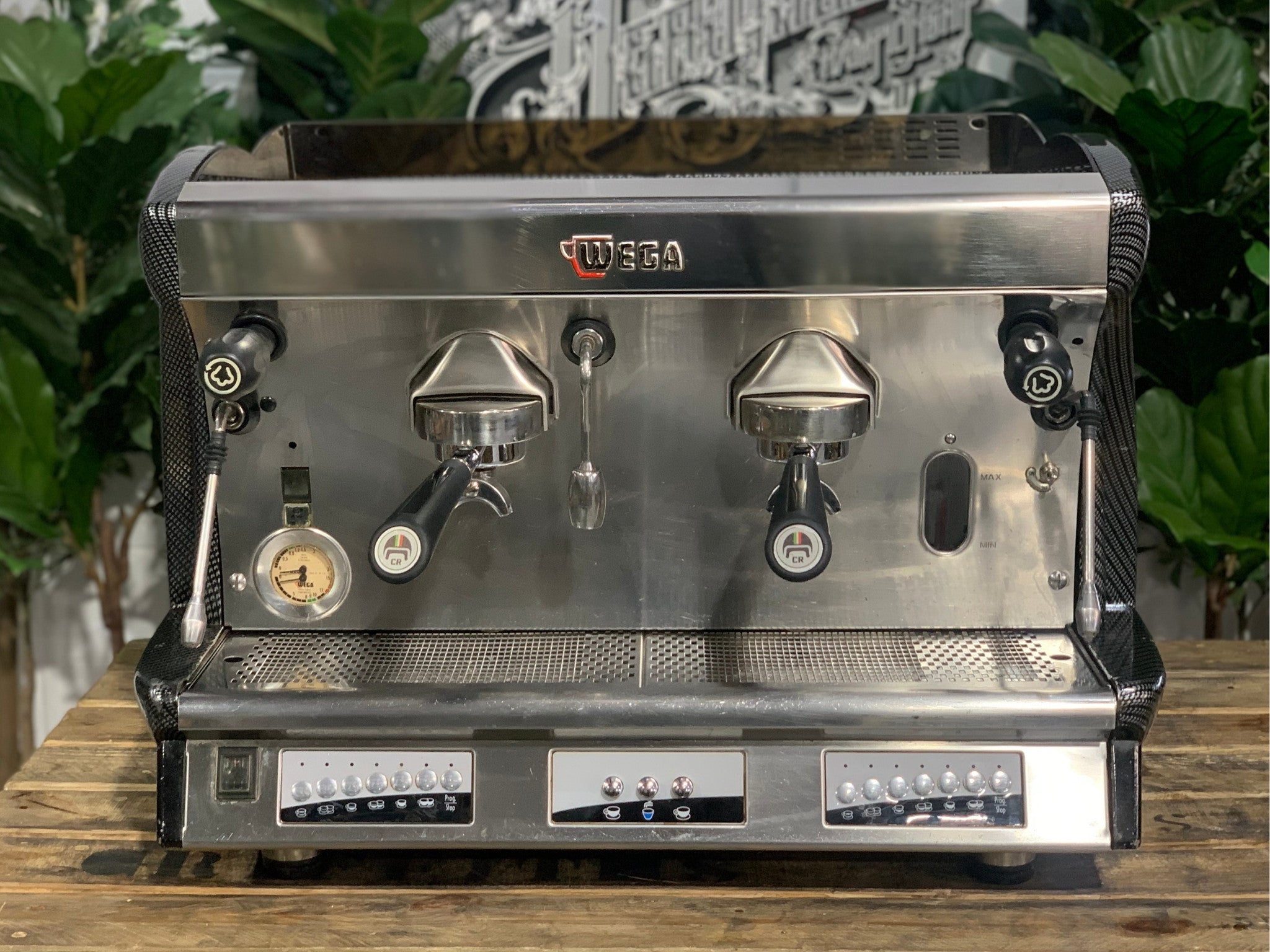 A Wega Vela 2 Group Espresso Coffee Machine with Carbon Fibre panels sitting on a wooden bench. There is greenery in the background. The image is taken from the front of the machine so that you can see the front of the machine, control panel, buttons and group heads.