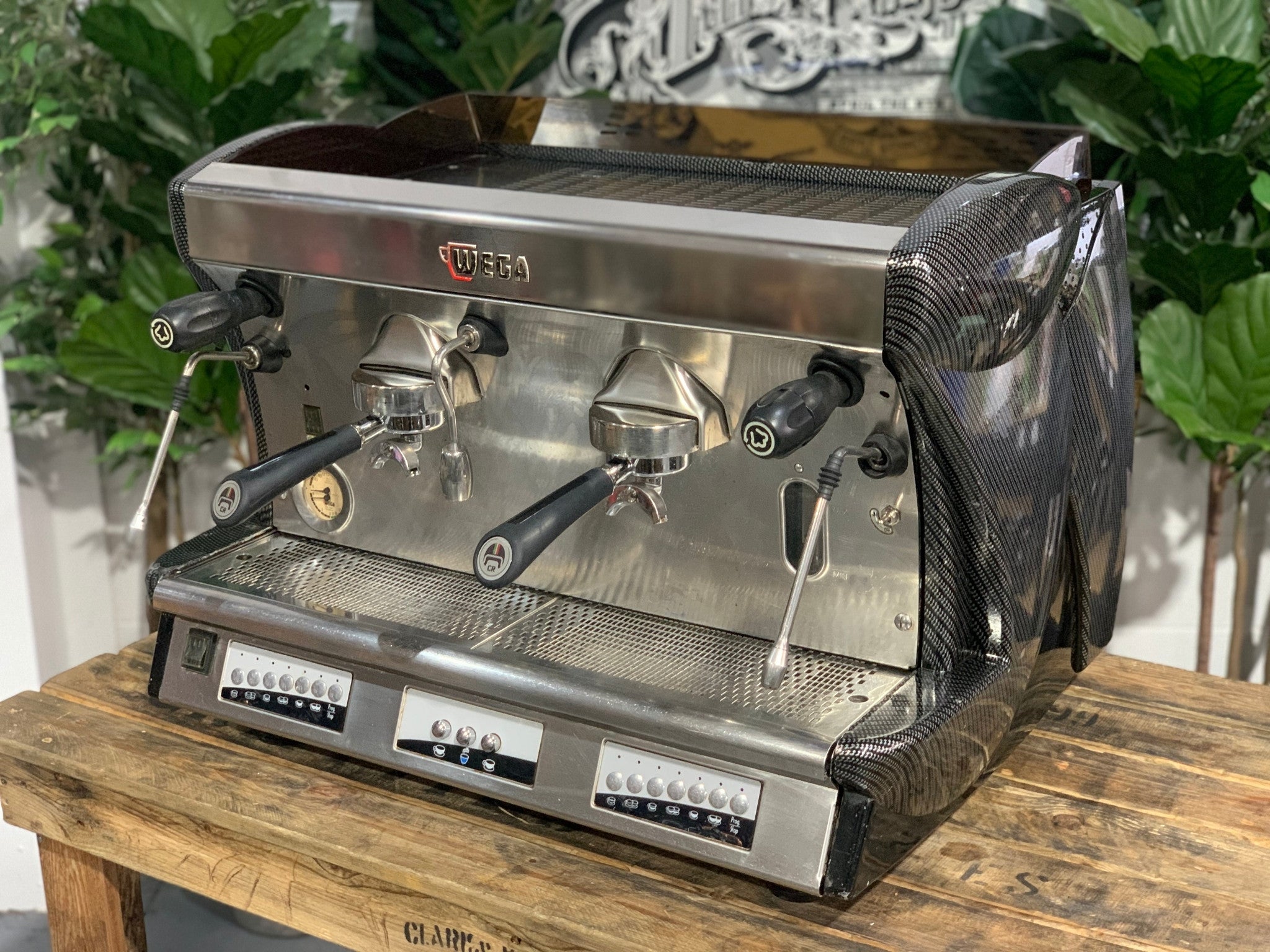 A Wega Vela 2 Group Espresso Coffee Machine with Carbon Fibre panels sitting on a wooden bench. There is greenery in the background. The image is taken from an angle so that the front as well as the side of the espresso machine is visible.