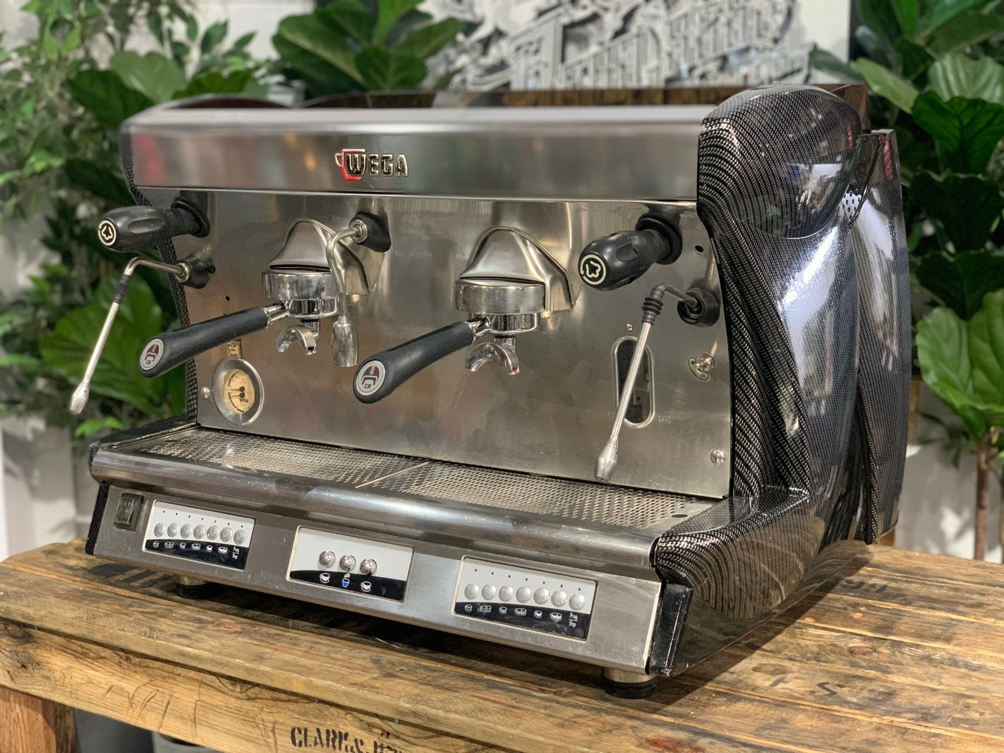 A Wega Vela 2 Group Espresso Coffee Machine with Carbon Fibre panels sitting on a wooden bench. There is greenery in the background. The image is taken from an angle so that the front as well as the side of the espresso machine is visible.
