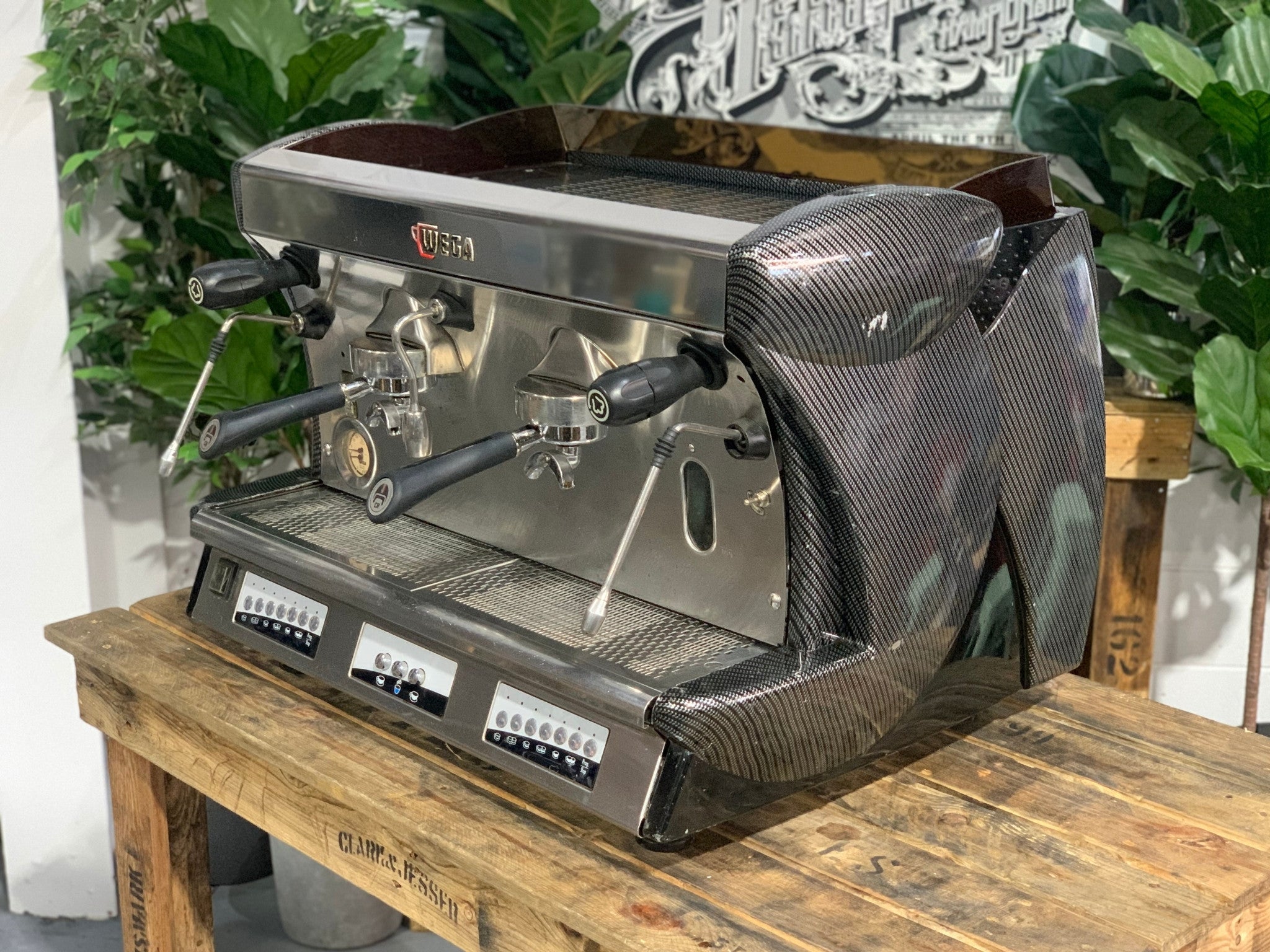 A Wega Vela 2 Group Espresso Coffee Machine with Carbon Fibre panels sitting on a wooden bench. There is greenery in the background. The image is taken from an angle so that the front as well as the side of the espresso machine is visible.