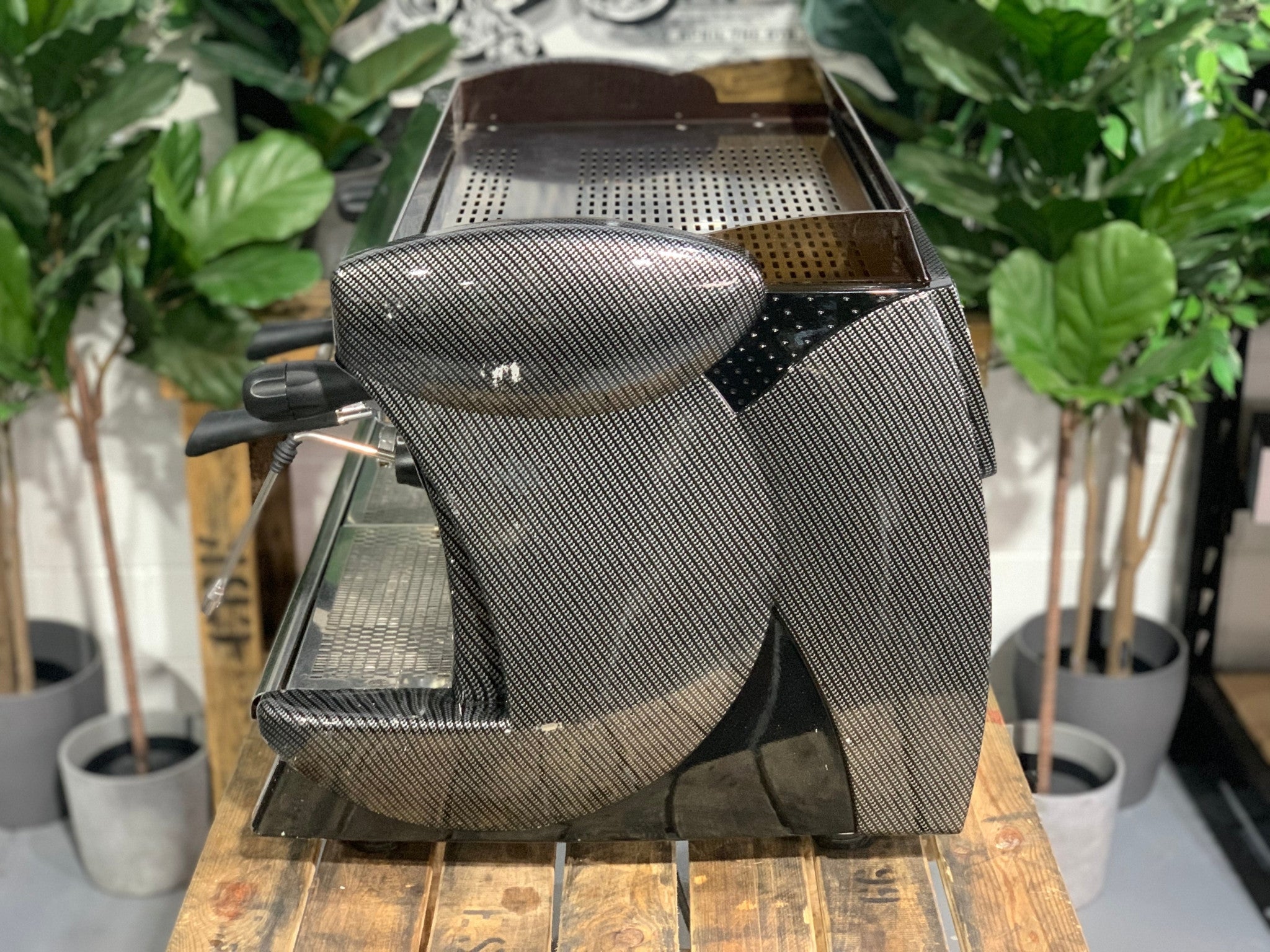 A Wega Vela 2 Group Espresso Coffee Machine with Carbon Fibre panels sitting on a wooden bench. There is greenery in the background. The image is taken from the side of the machine.