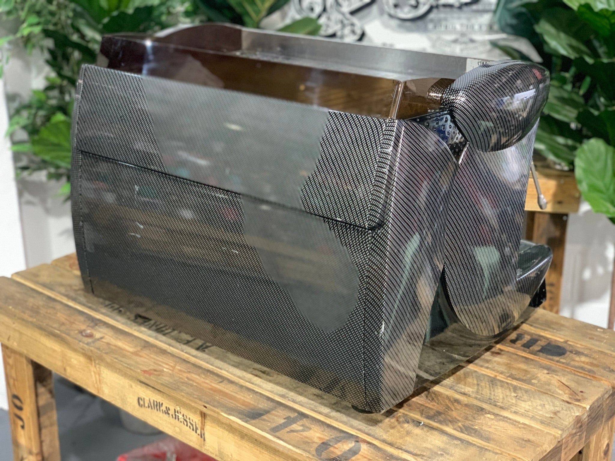 A Wega Vela 2 Group Espresso Coffee Machine with Carbon Fibre panels sitting on a wooden bench. There is greenery in the background. The image is taken from behind the machine and the machine is sitting on an angle so that you can see the back of the machine and the side.