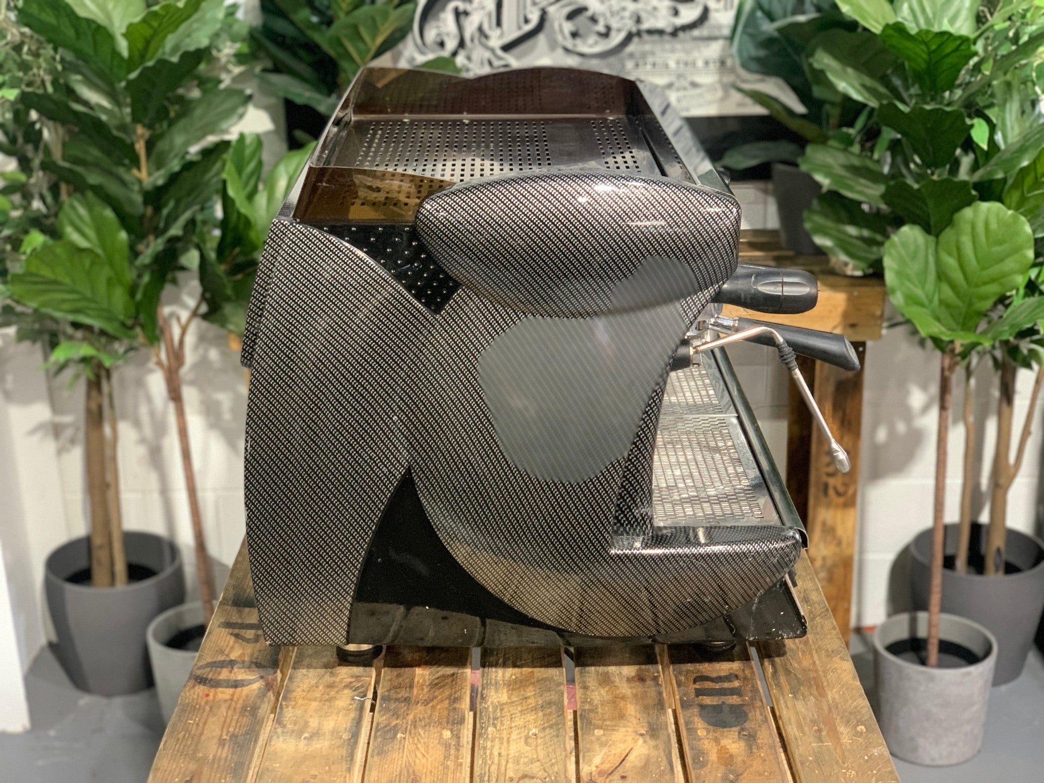 A Wega Vela 2 Group Espresso Coffee Machine with Carbon Fibre panels sitting on a wooden bench. There is greenery in the background. The image is taken from the side of the machine.