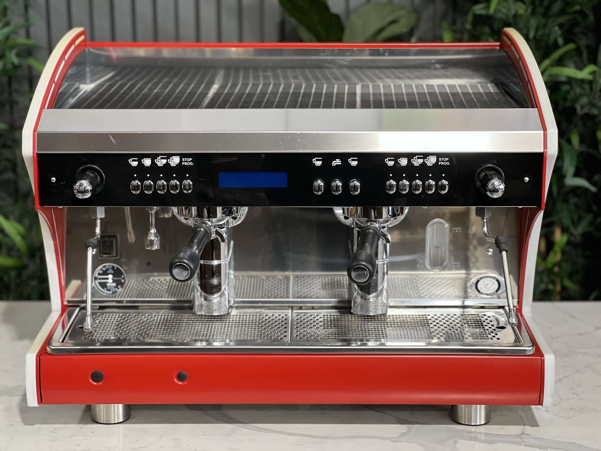 Wega Polaris Tron 2 Group Espresso Coffee Machine with red finish is sitting on a marble bench. There is greenery in the background. The image is taken from the front of the machine so that you can see the front of the machine, control panel, buttons and group heads.