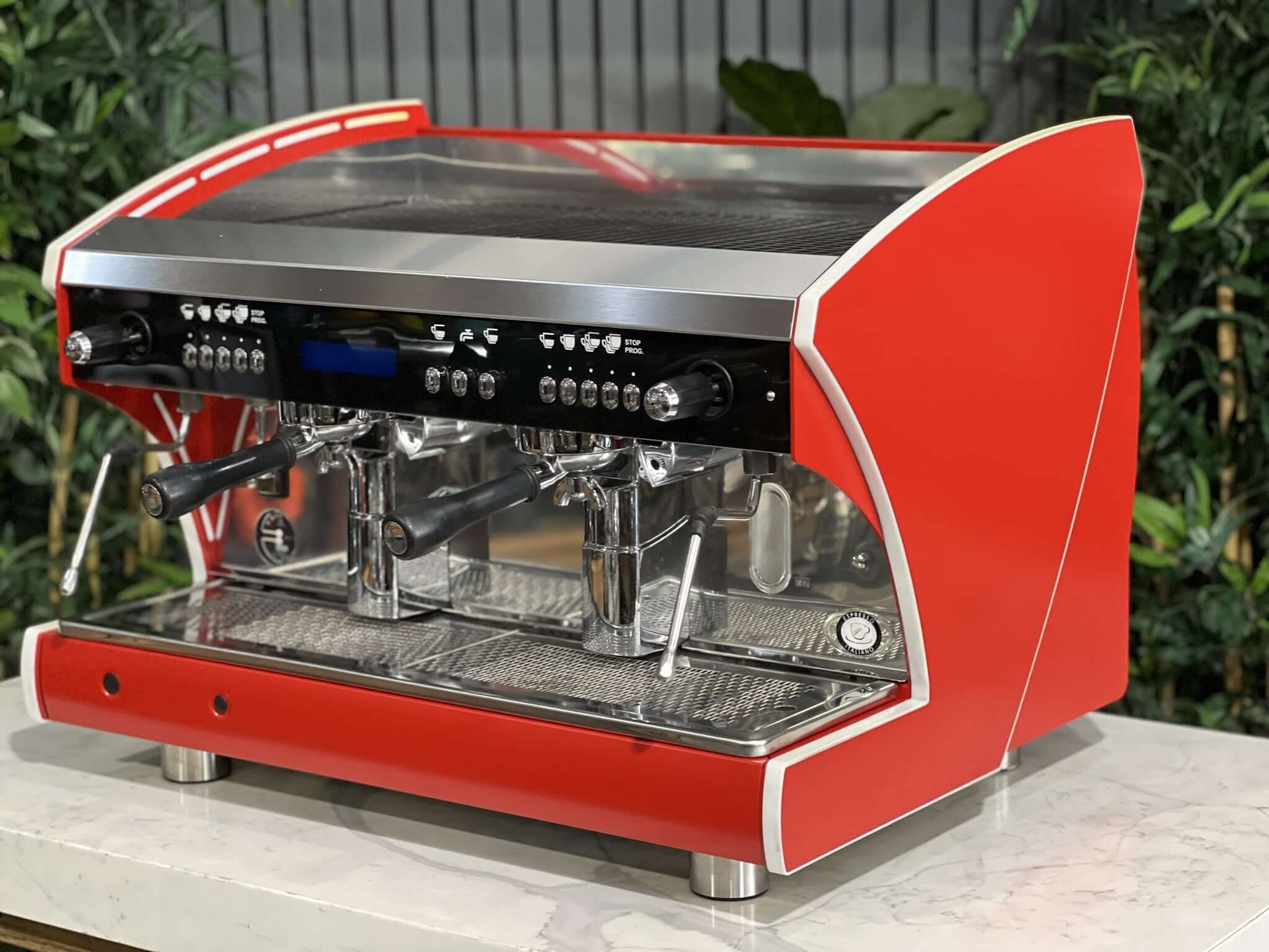 Wega Polaris Tron 2 Group Espresso Coffee Machine with red finish  is sitting on a marble bench. There is greenery in the background. The image is taken from the front of the machine and the machine is sitting on an angle so that you can see the front of the machine and the side.