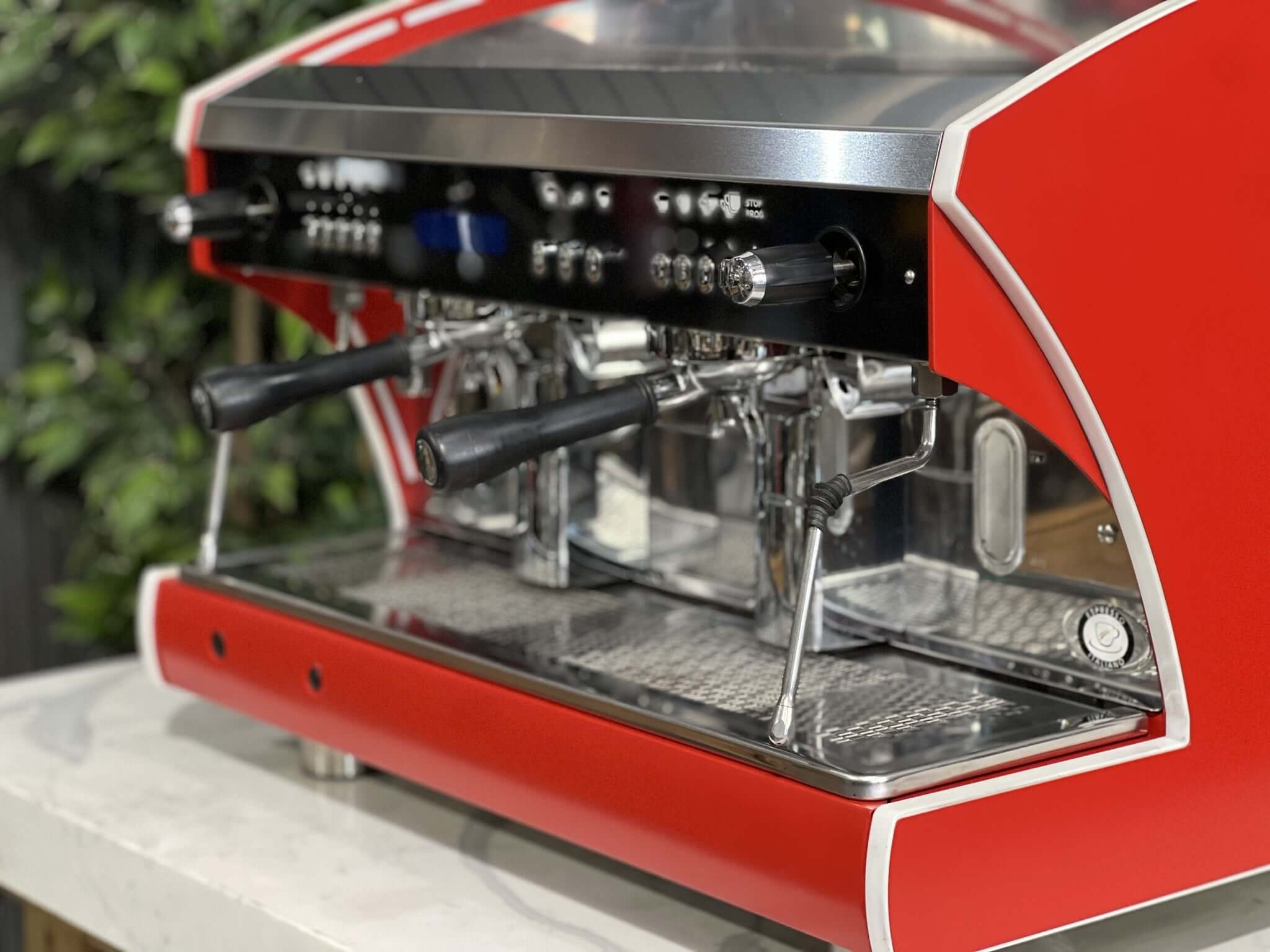 Wega Polaris Tron 2 Group Espresso Coffee Machine with red finish is sitting on a marble bench. There is greenery in the background. The image is a close up image taken from the front of the machine and the machine is sitting on an angle so that you can see the front of the machine, the controls including buttons, group handles and logo.