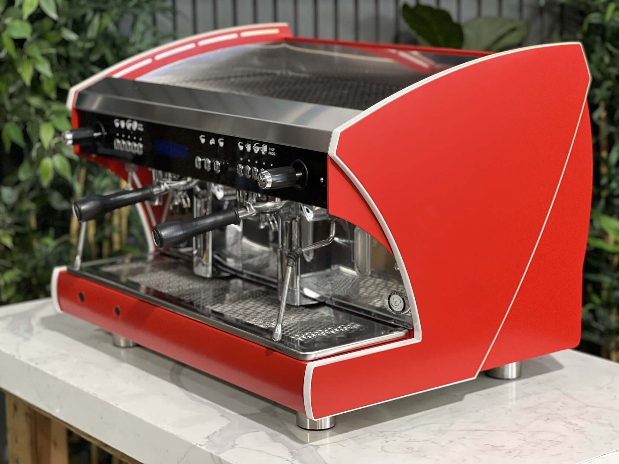 Wega Polaris Tron 2 Group Espresso Coffee Machine with red finish is sitting on a marble bench. There is greenery in the background. The image is taken from the front of the machine and the machine is sitting on an angle so that you can see the front of the machine and the side.