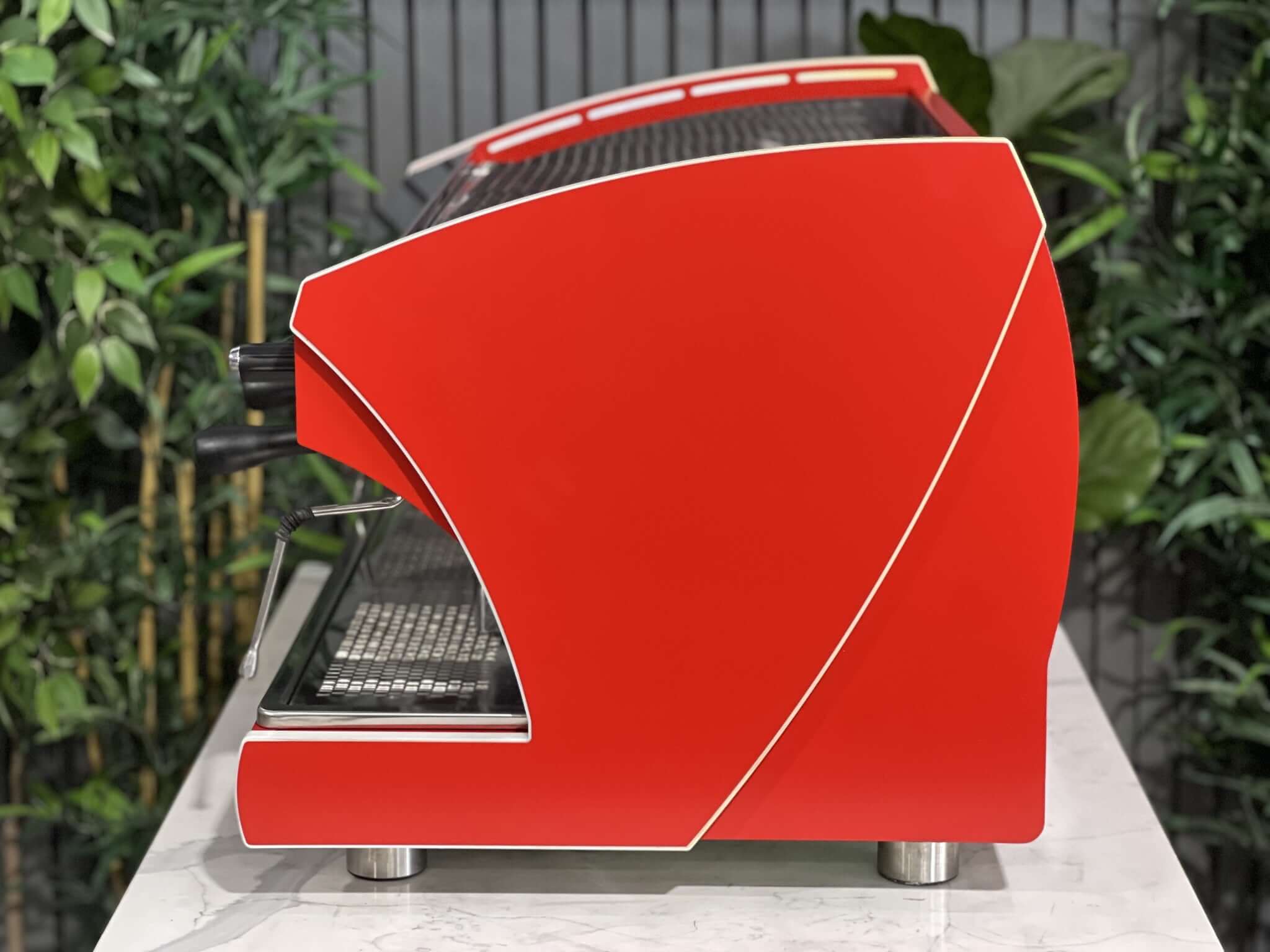 Wega Polaris Tron 2 Group Espresso Coffee Machine with red finish is sitting on a marble bench. There is greenery in the background. The image is taken from the side of the machine.