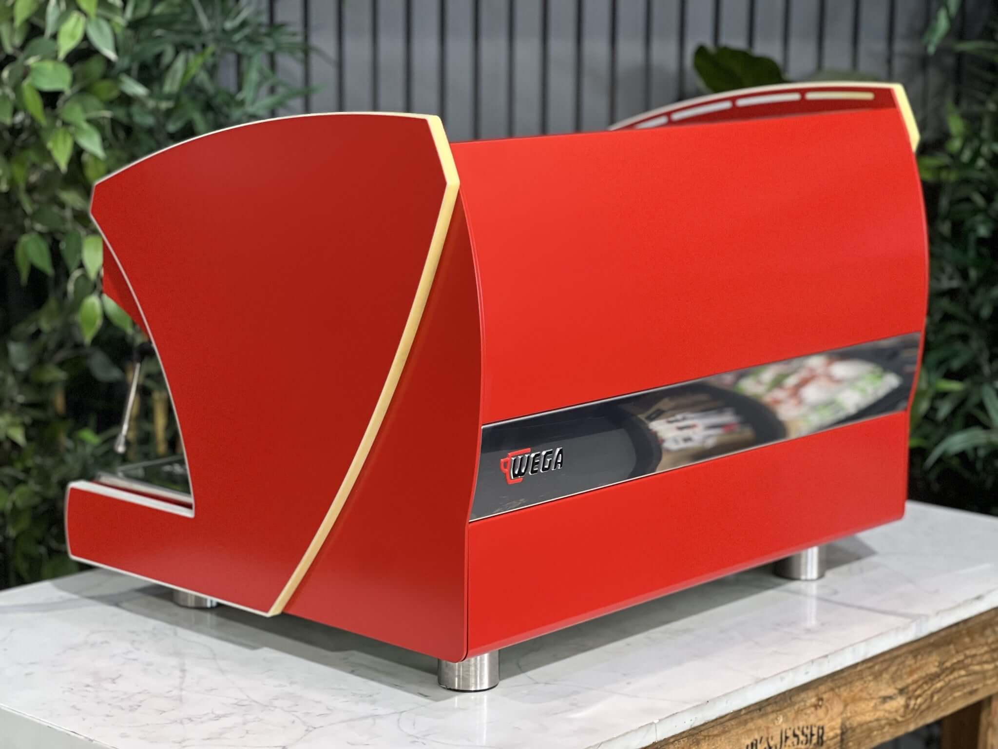 Wega Polaris Tron 2 Group Espresso Coffee Machine with red finish sitting on a marble bench. There is greenery in the background. The image is taken from behind the machine and the machine is sitting on an angle so that you can see the back of the machine and the side.