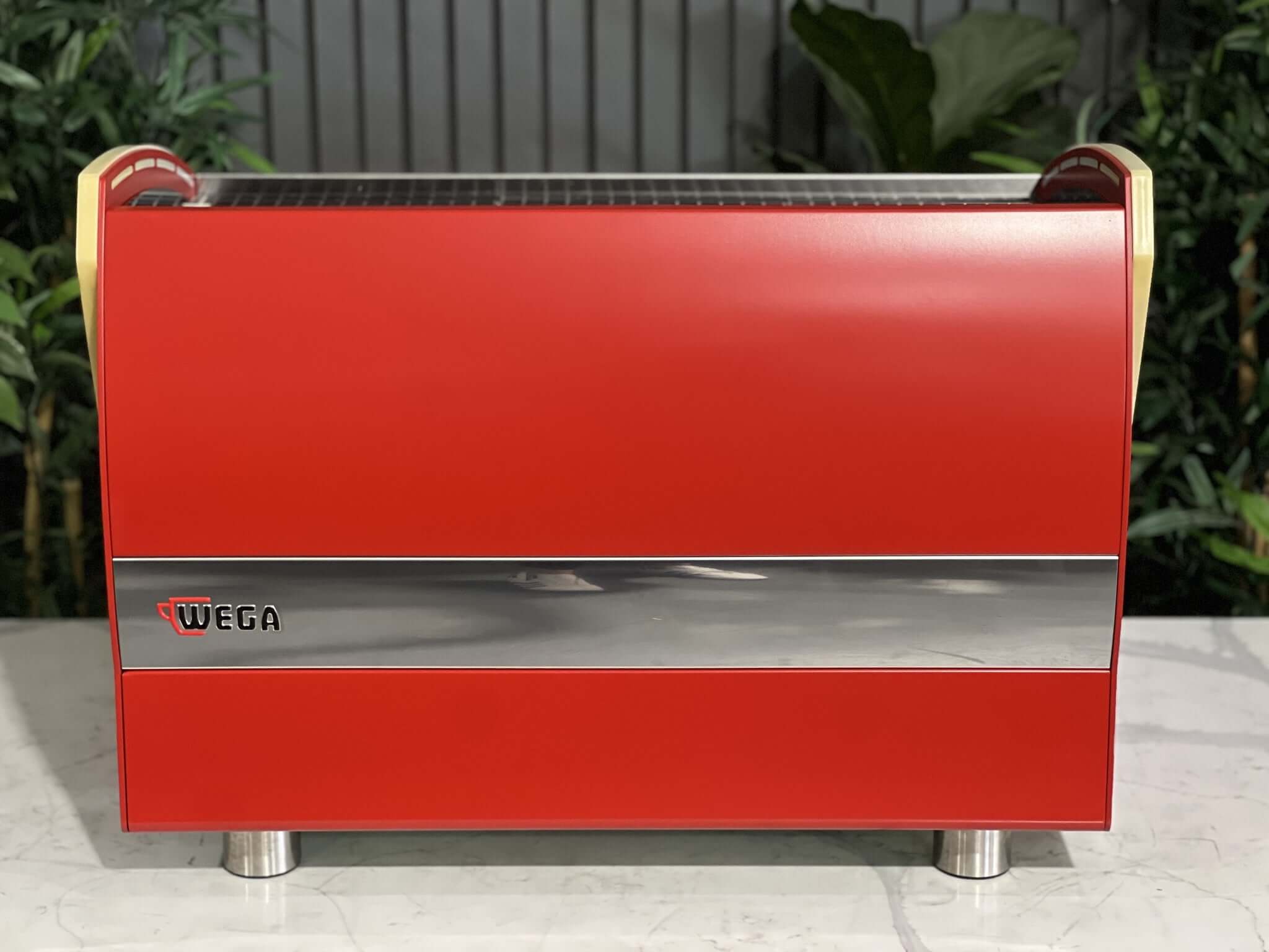 Wega Polaris Tron 2 Group Espresso Coffee Machine with red finish is sitting on a marble bench. There is greenery in the background. The image is taken from behind the machine so the back is visible.