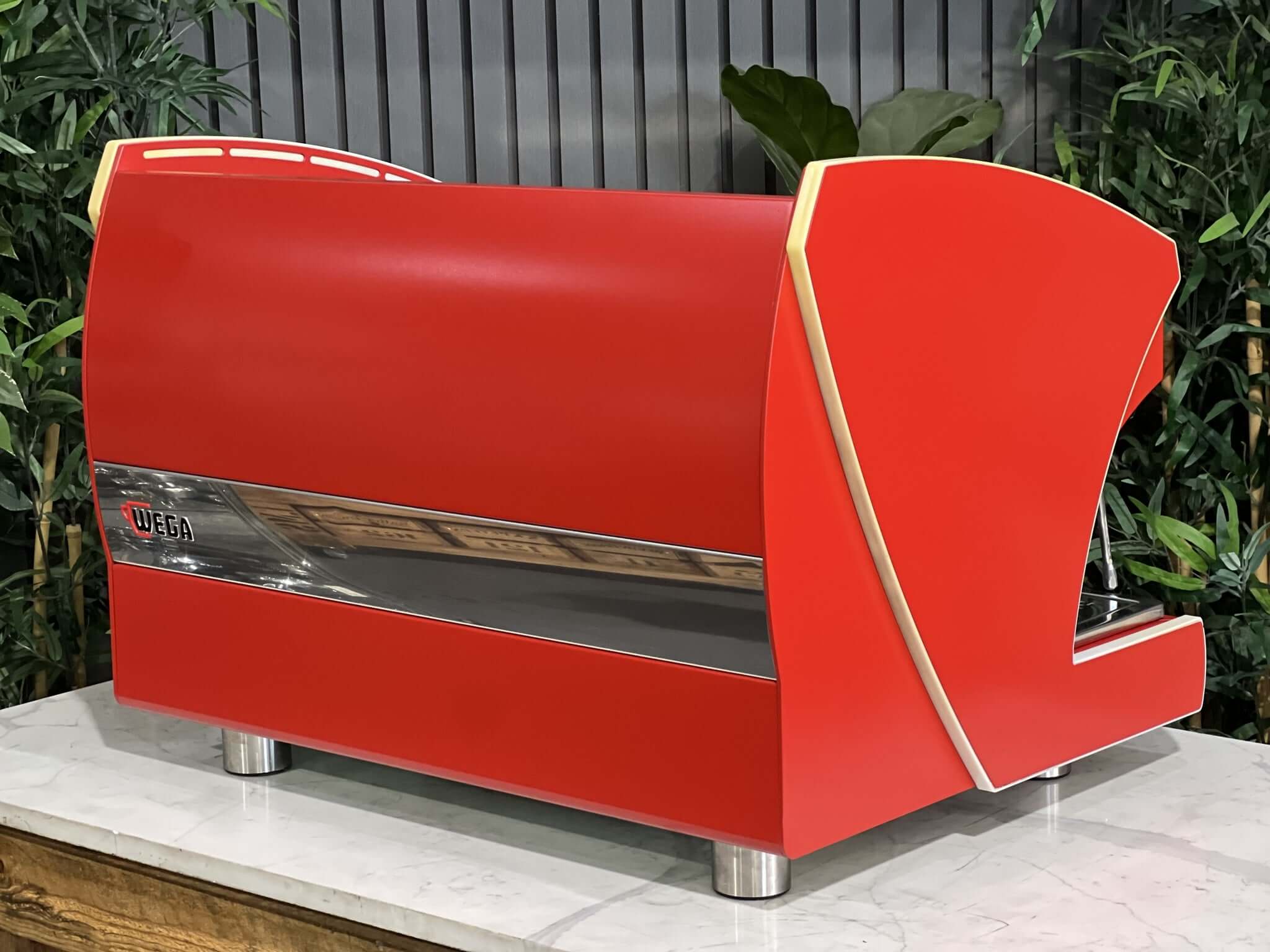 Wega Polaris Tron 2 Group Espresso Coffee Machine with red finish is sitting on a marble bench. There is greenery in the background. The image is taken from behind the machine and the machine is sitting on an angle so that you can see the back of the machine and the side.