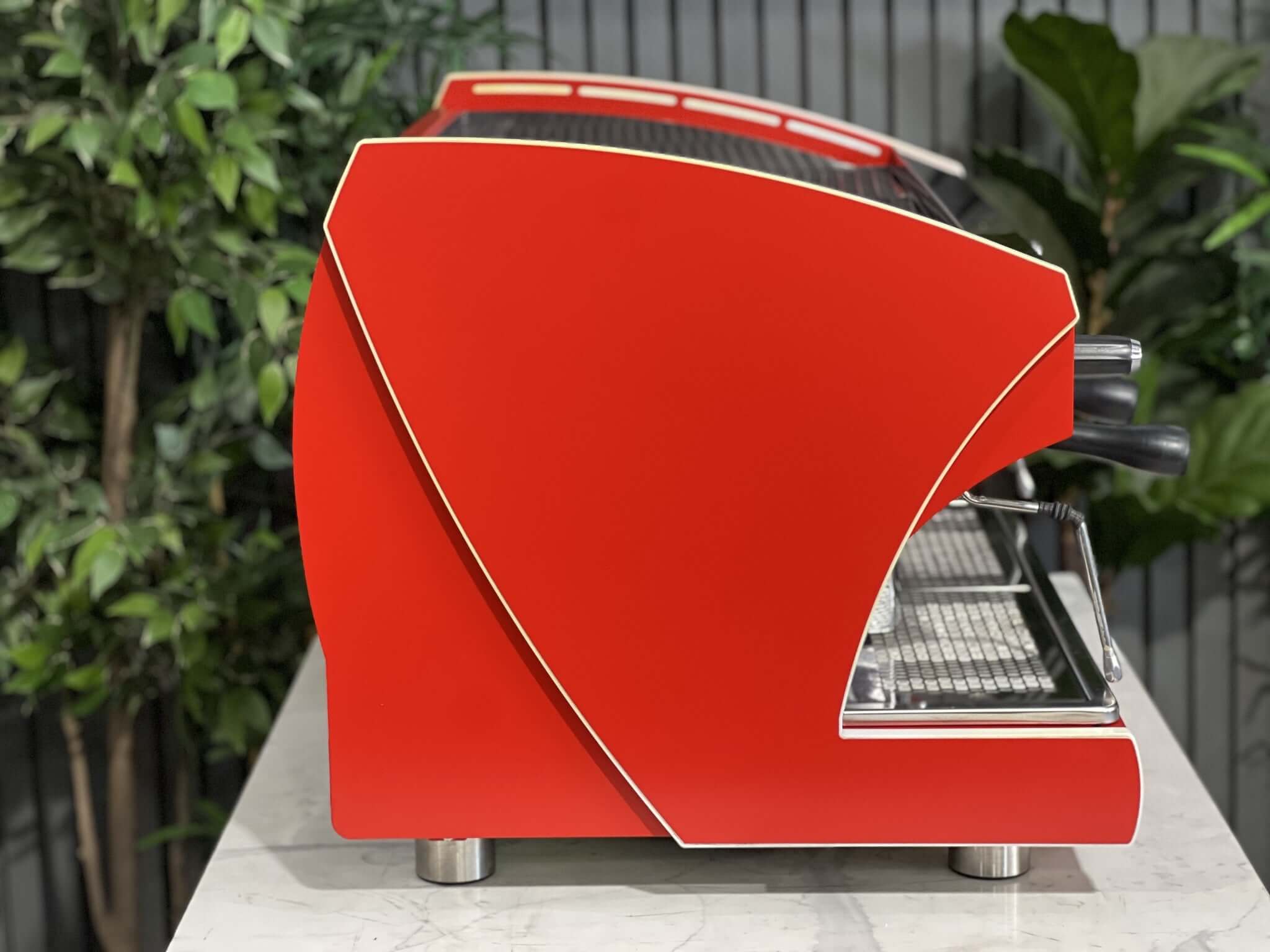 Wega Polaris Tron 2 Group Espresso Coffee Machine with red finish is sitting on a marble bench. There is greenery in the background. The image is taken from the side of the machine.
