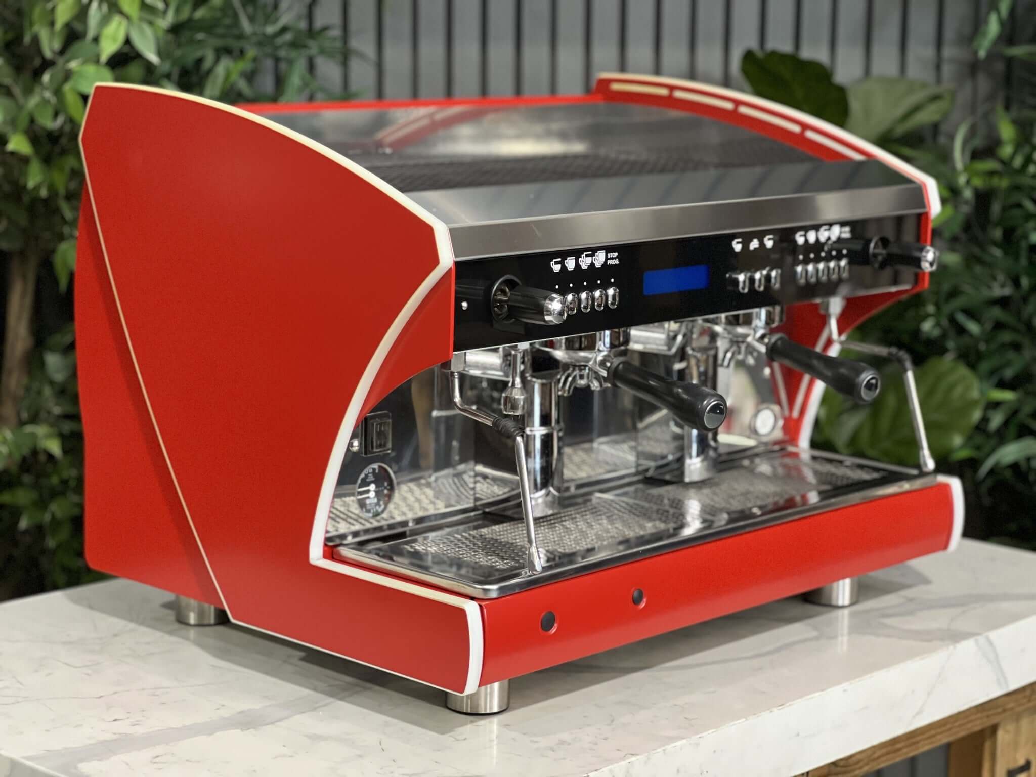 Wega Polaris Tron 2 Group Espresso Coffee Machine with red finish is sitting on a marble bench. There is greenery in the background. The image is taken from the front of the machine and the machine is sitting on an angle so that you can see the front of the machine and the side.