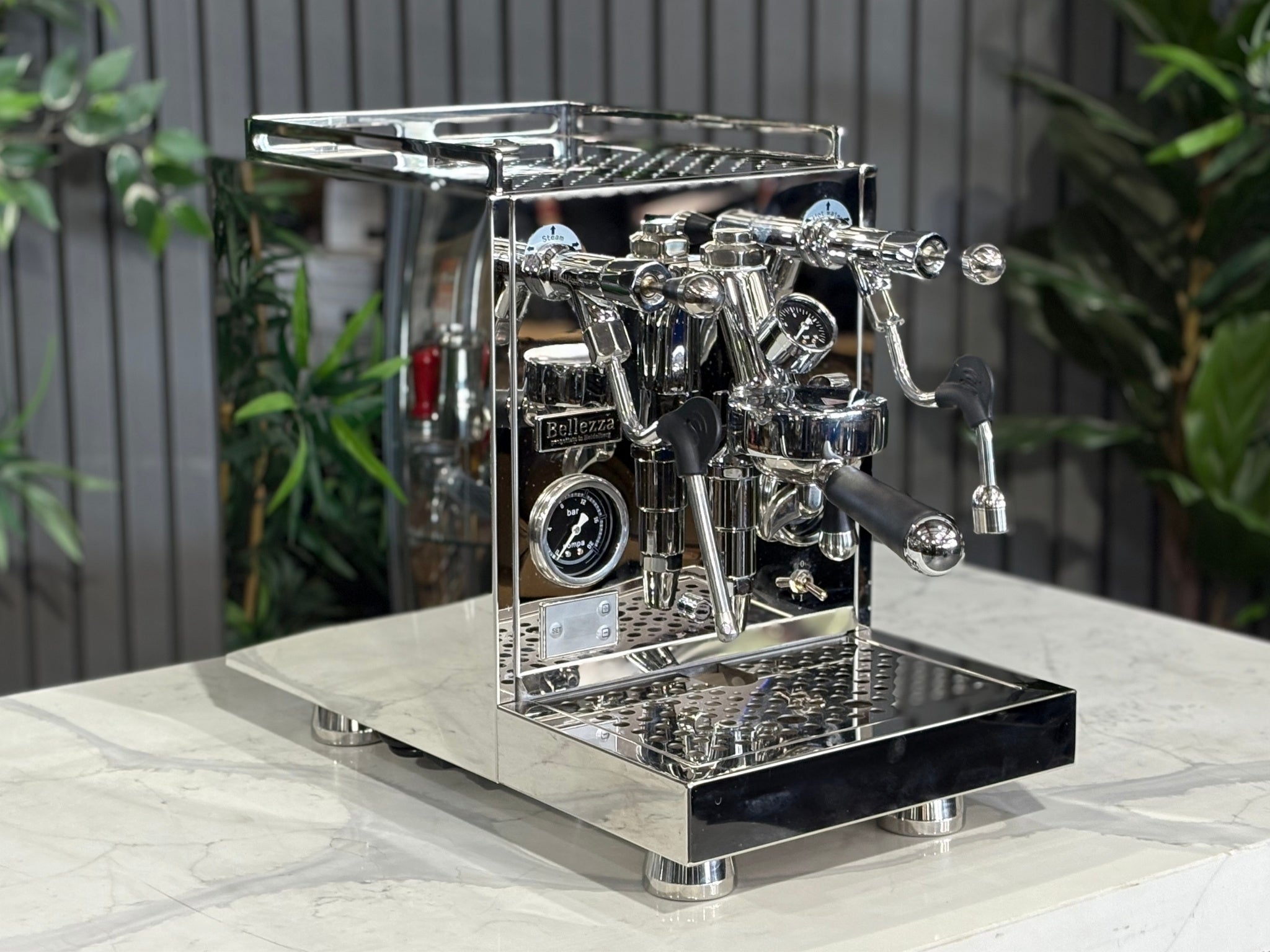 A Bellezza Inizio R 1 Group Coffee Machine with Stainless finish  is sitting on a marble bench. There is greenery in the background. The image is taken from the front of the machine and the machine is sitting on an angle so that you can see the front of the machine and the side. 