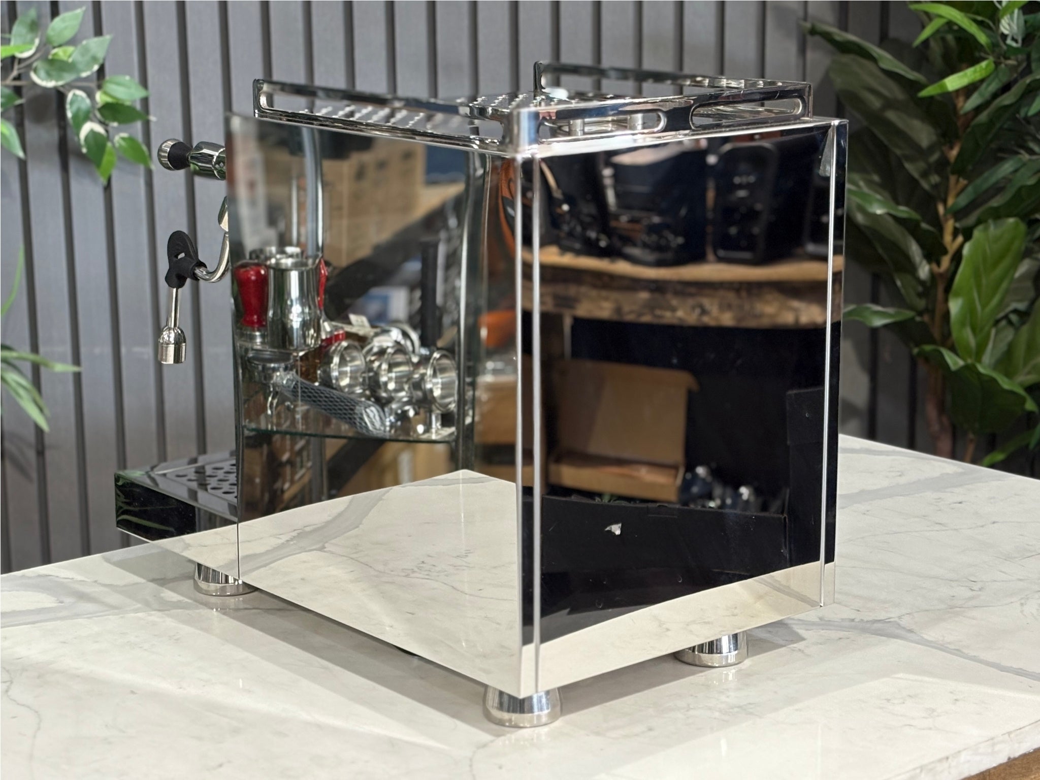 A Bellezza Inizio R 1 Group Coffee Machine with Stainless finish sitting on a marble bench. There is greenery in the background. The image is taken from behind the machine and the machine is sitting on an angle so that you can see the back of the machine and the side.