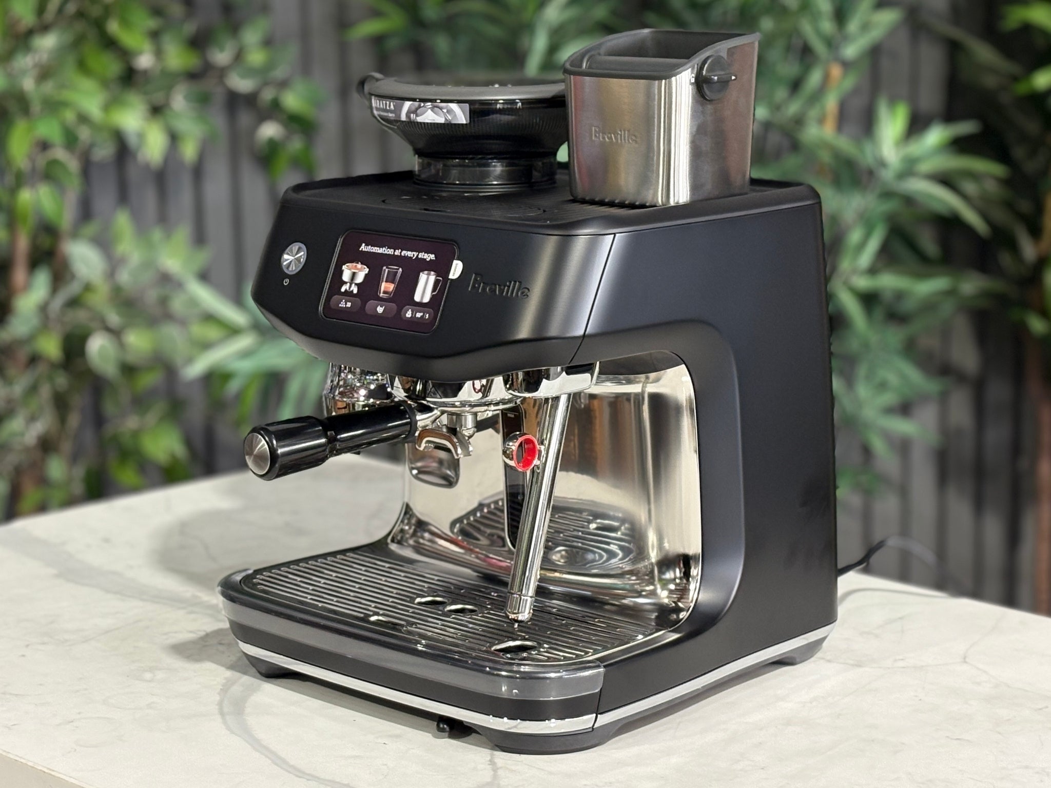 Breville Oracle Jet 1 Group Coffee Machine Black - New, angled view on a marble bench with greenery in the background, showing the black body, touch screen display, milk steamer, and hopper.