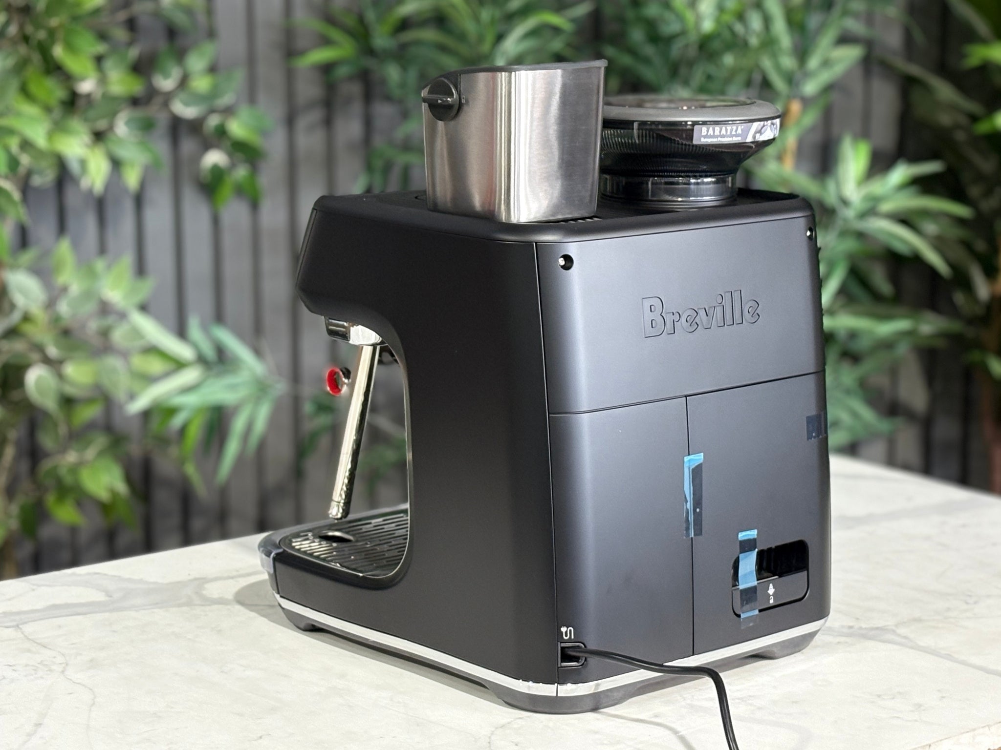 Breville Oracle Jet 1 Group Coffee Machine Black - New, angled rear view on a marble bench with greenery, showing the black body, water reservoir area, and overall sleek design.