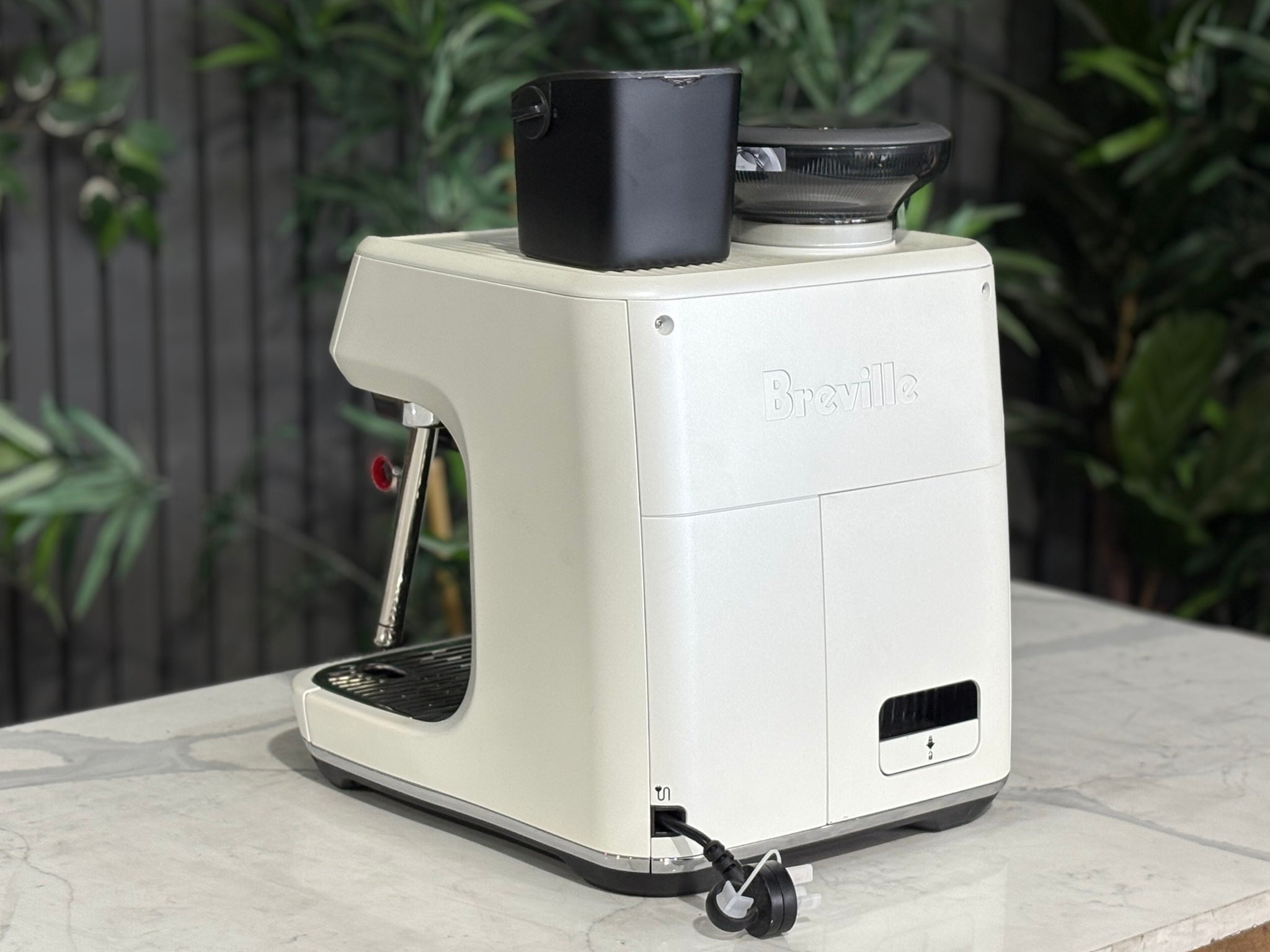 Breville Oracle Jet 1 Group Coffee Machine White - New, angled rear view on a marble bench with greenery, showing the white espresso machine body, water reservoir area, and sleek design.