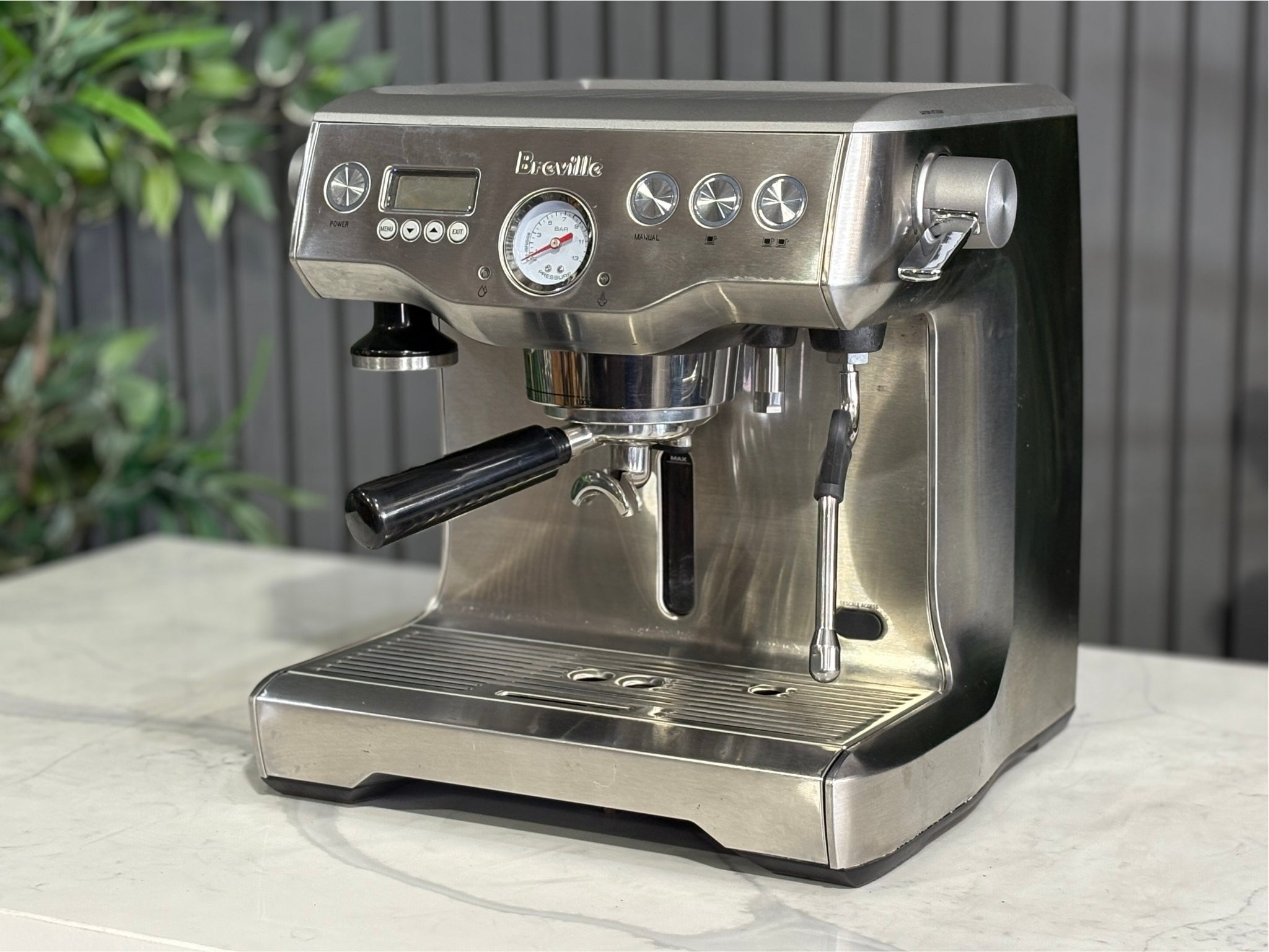 A Breville The Dual Boiler BES920 Espresso Coffee Machine with Stainless Steel finish is sitting on a marble bench. There is greenery in the background. The image is taken from the front of the machine and the machine is sitting on an angle so that you can see the front of the machine and the side. 