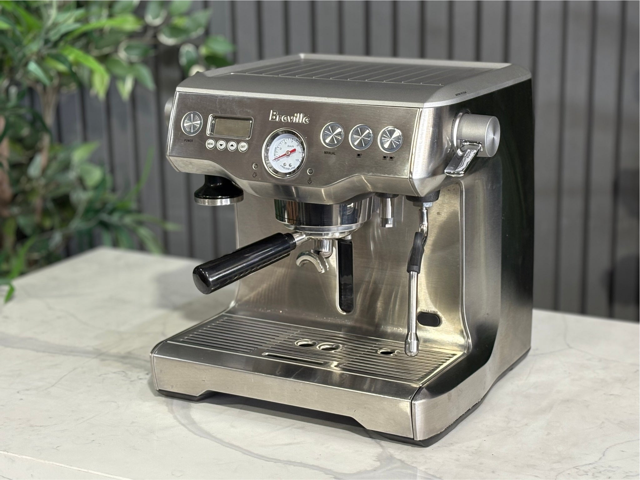 A Breville The Dual Boiler BES920 Espresso Coffee Machine with Stainless Steel finish  is sitting on a marble bench. There is greenery in the background. The image is taken from the front of the machine and the machine is sitting on an angle so that you can see the front of the machine and the side.