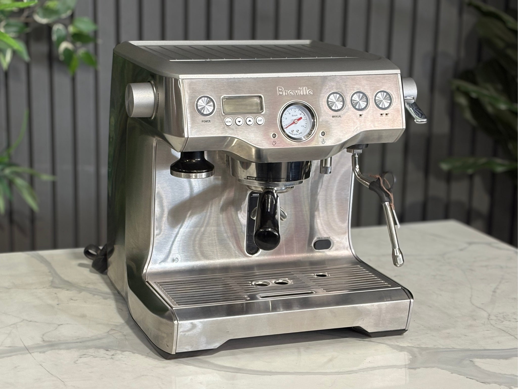 A Breville The Dual Boiler BES920 Espresso Coffee Machine with Stainless Steel finish is sitting on a marble bench. There is greenery in the background. The image is taken from the front of the machine and the machine is sitting on an angle so that you can see the front of the machine and the side. 