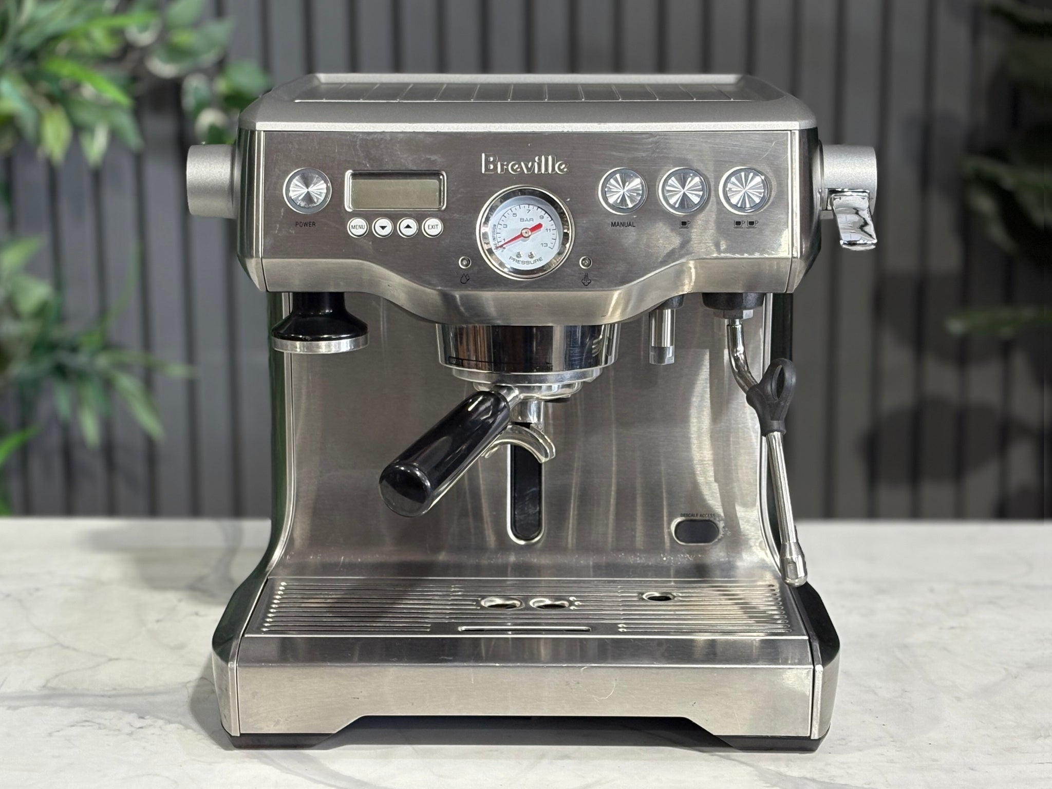 A Breville The Dual Boiler BES920 Espresso Coffee Machine with Stainless Steel finish sitting on a marble bench. There is greenery in the background. The image is taken from the front of the machine so that you can see the front of the machine, control panel, buttons and group heads.
