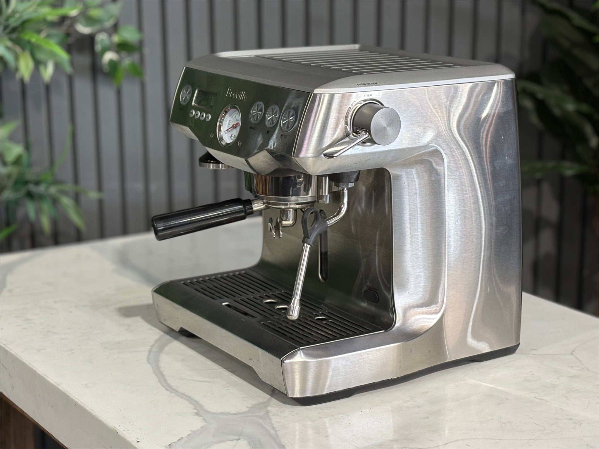 A Breville The Dual Boiler BES920 Espresso Coffee Machine with Stainless Steel finish  is sitting on a marble bench. There is greenery in the background. The image is taken from the front of the machine and the machine is sitting on an angle so that you can see the front of the machine and the side.