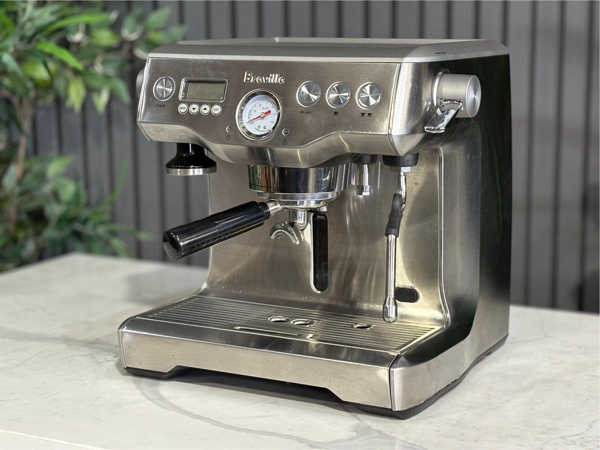 A Breville The Dual Boiler BES920 Espresso Coffee Machine with Stainless Steel finish  is sitting on a marble bench. There is greenery in the background. The image is taken from the front of the machine and the machine is sitting on an angle so that you can see the front of the machine and the side.