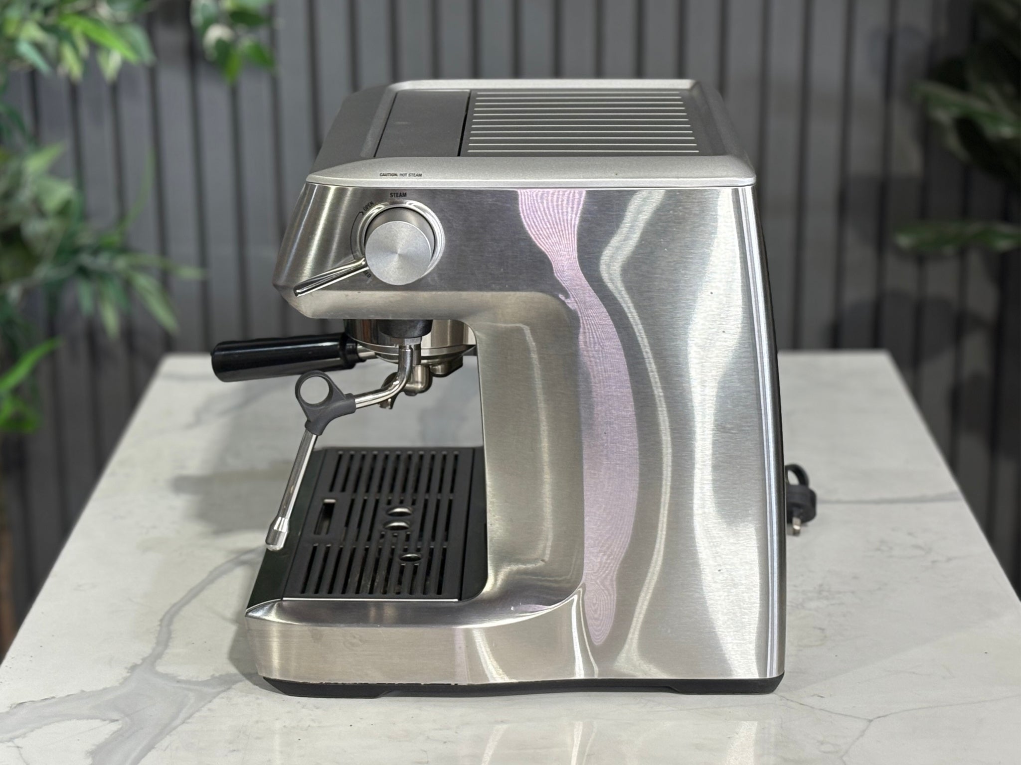 A Breville The Dual Boiler BES920 Espresso Coffee Machine with Stainless Steel finish sitting on a marble bench. There is greenery in the background. The image is taken from the side of the machine.
