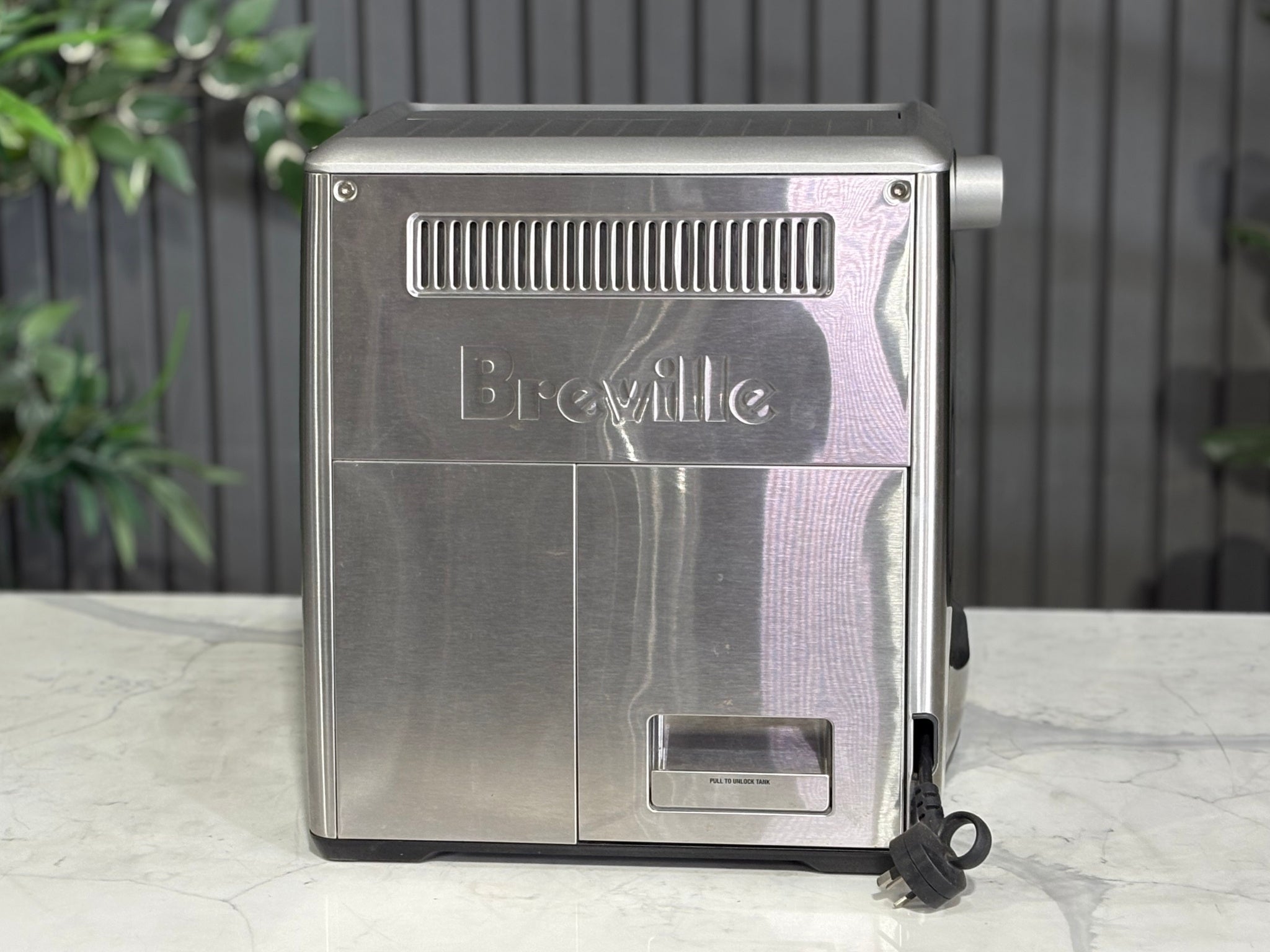 A Breville The Dual Boiler BES920 Espresso Coffee Machine with Stainless Steel finish is sitting on a marble bench. There is greenery in the background. The image is taken from behind the machine so the back is visible. 