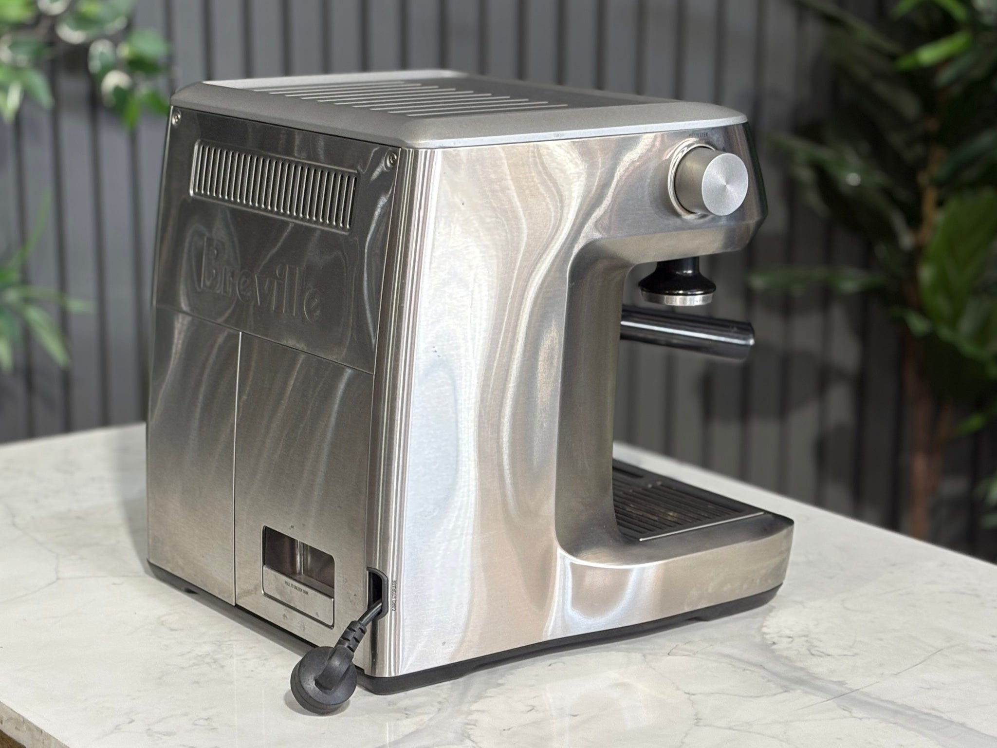 A Breville The Dual Boiler BES920 Espresso Coffee Machine with Stainless Steel finish sitting on a marble bench. There is greenery in the background. The image is taken from behind the machine and the machine is sitting on an angle so that you can see the back of the machine and the side.