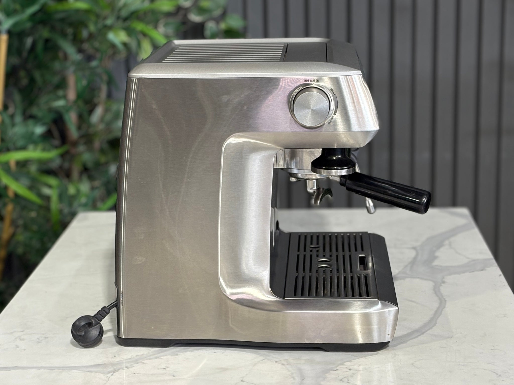 A Breville The Dual Boiler BES920 Espresso Coffee Machine with Stainless Steel finish sitting on a marble bench. There is greenery in the background. The image is taken from the side of the machine.