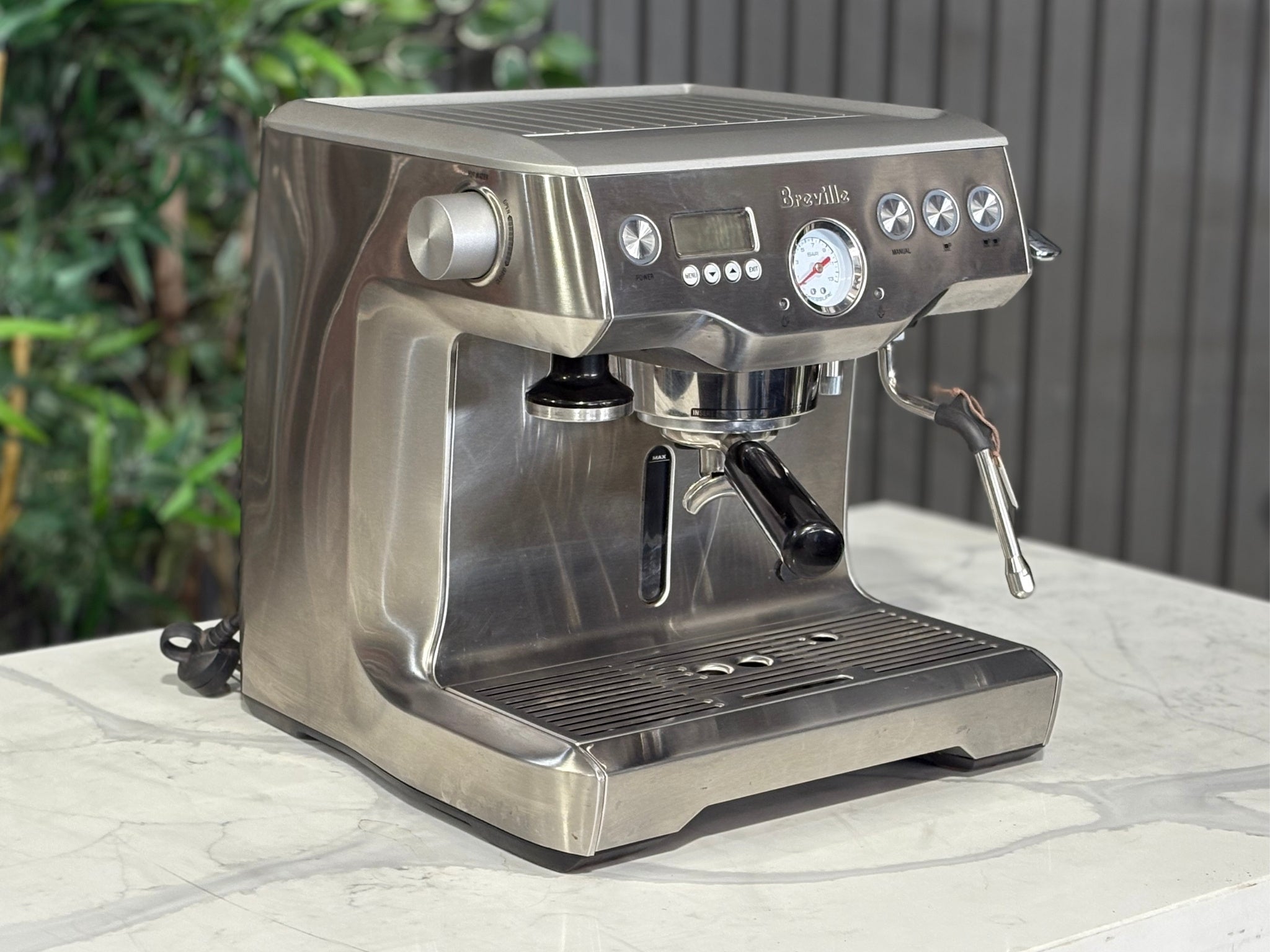 A Breville The Dual Boiler BES920 Espresso Coffee Machine with Stainless Steel finish is sitting on a marble bench. There is greenery in the background. The image is taken from the front of the machine and the machine is sitting on an angle so that you can see the front of the machine and the side. 