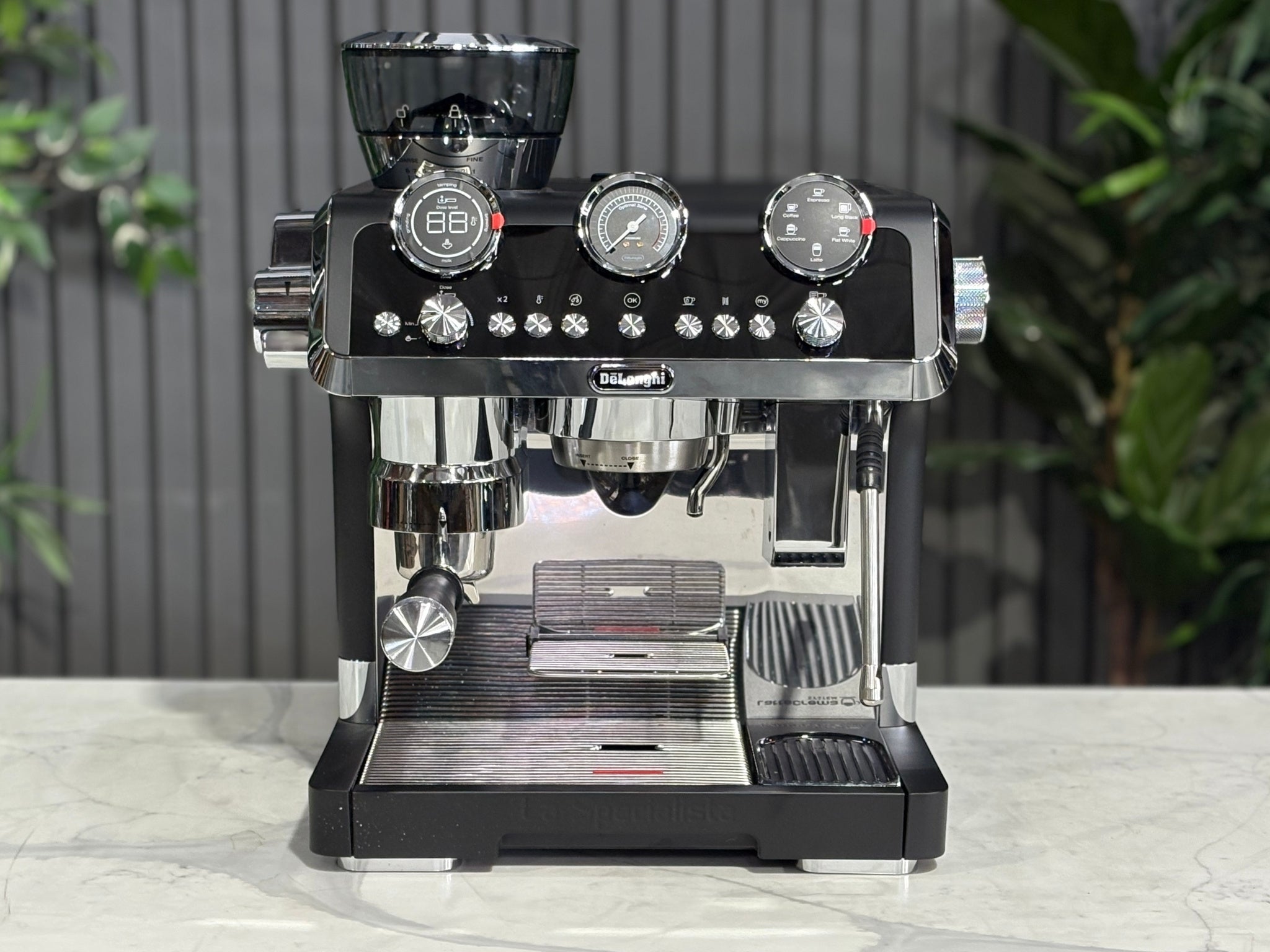 A DeLonghi La Specialista Maestro 1 Group Coffee Machine in Black sitting on a marble bench. There is greenery in the background. The image is taken from the front of the machine so that you can see the front of the machine, control panel, buttons and group head.