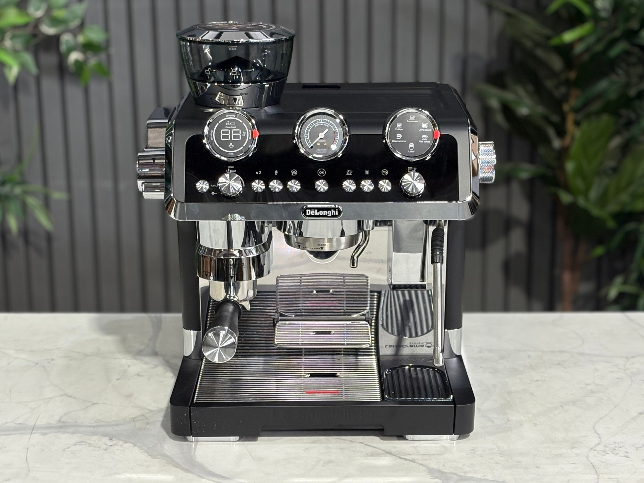 A DeLonghi La Specialista Maestro 1 Group Coffee Machine in Blacksitting on a marble bench. There is greenery in the background. The photo is taken from a high front angle, clearly showcasing both the front panel and the top of the machine.