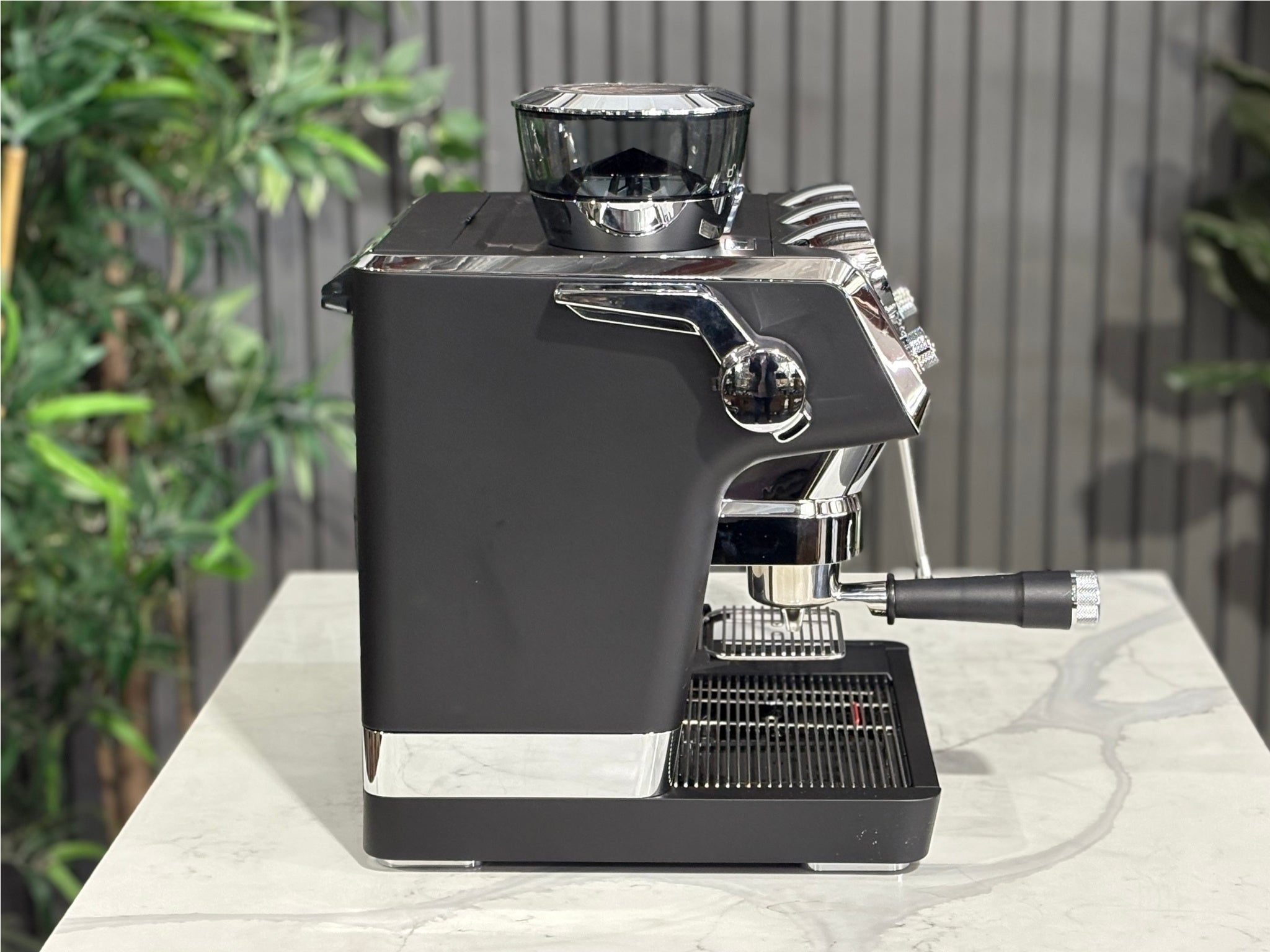 A DeLonghi La Specialista Maestro 1 Group Coffee Machine in Black sitting on a marble bench. There is greenery in the background. The image is taken from the side of the machine.