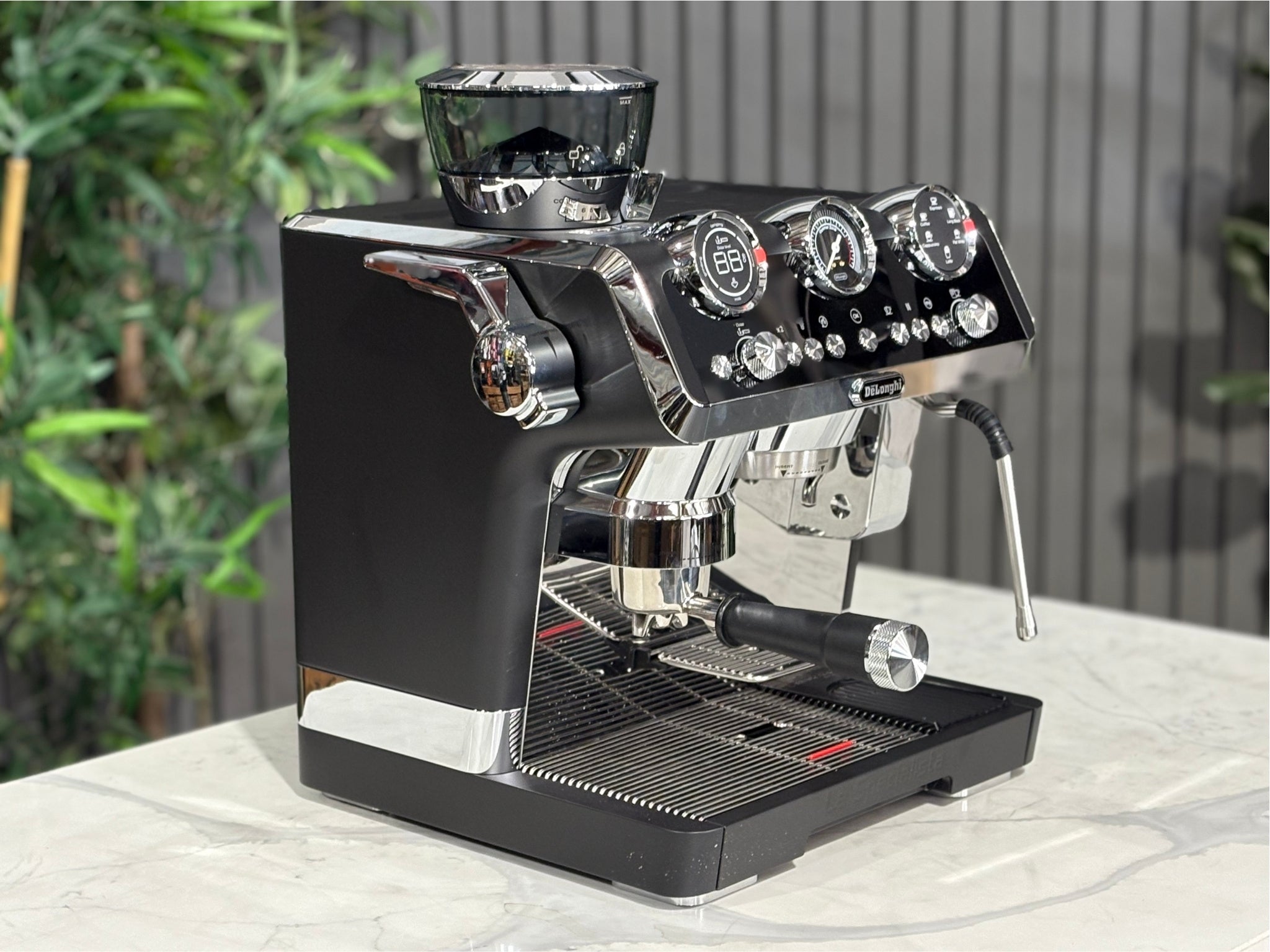 A DeLonghi La Specialista Maestro 1 Group Coffee Machine in Black  is sitting on a marble bench. There is greenery in the background. The image is taken from the front of the machine and the machine is sitting on an angle so that you can see the front of the machine and the side. 