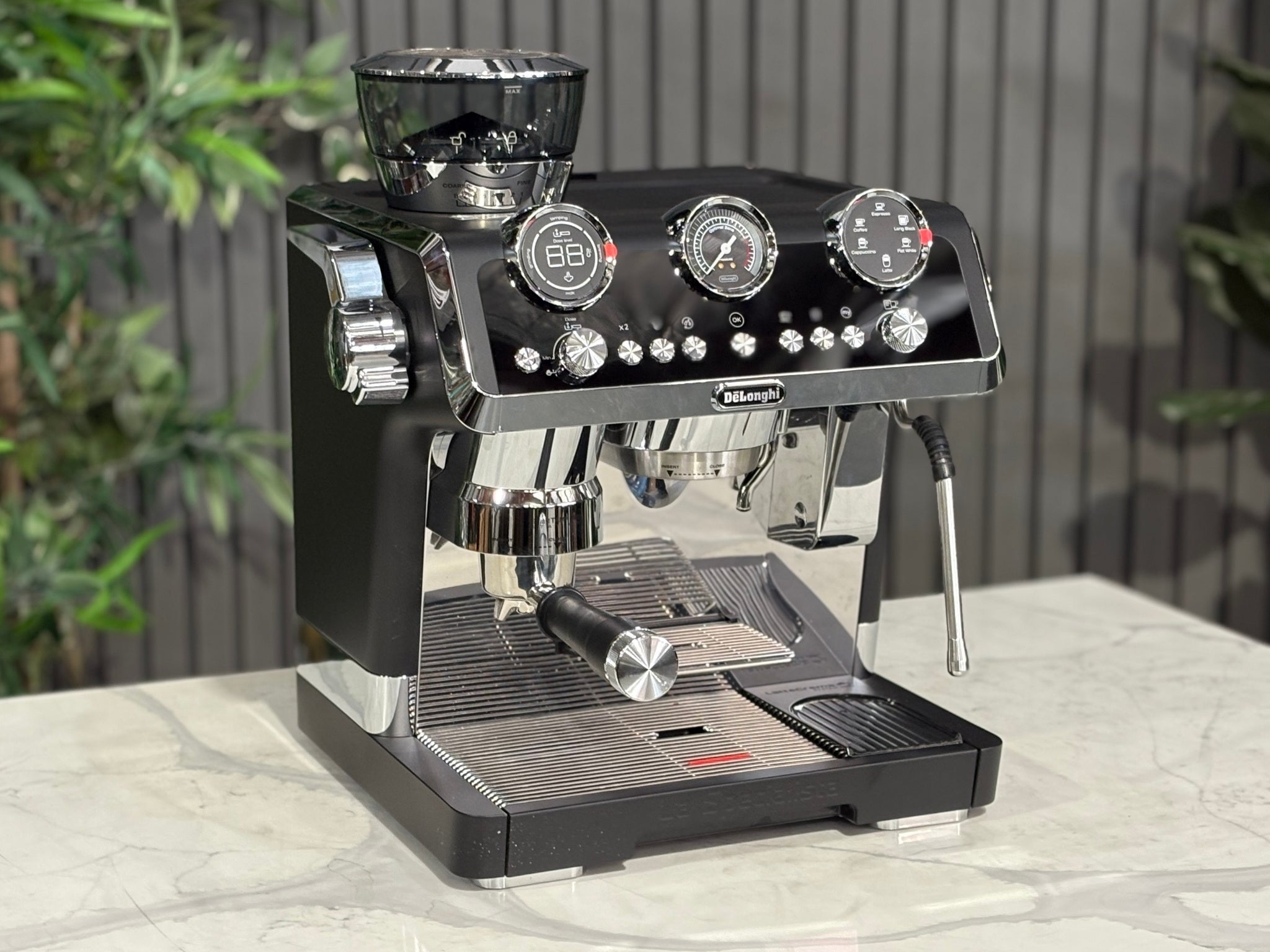 A DeLonghi La Specialista Maestro 1 Group Coffee Machine in Black  is sitting on a marble bench. There is greenery in the background. The image is taken from the front of the machine and the machine is sitting on an angle so that you can see the front of the machine and the side. 