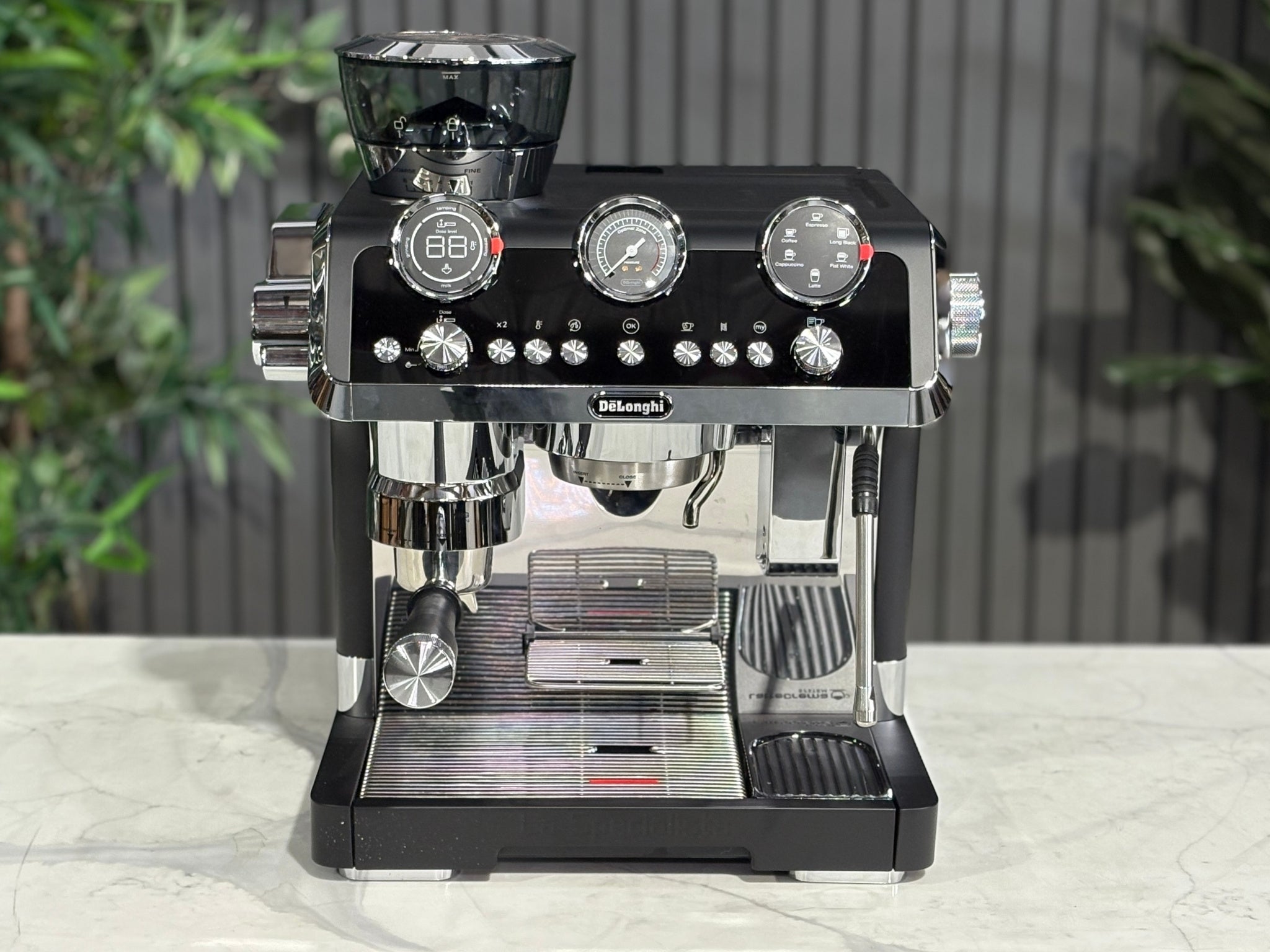 A DeLonghi La Specialista Maestro 1 Group Coffee Machine in Black sitting on a marble bench. There is greenery in the background. The photo is taken from a high front angle, clearly showcasing both the front panel and the top of the machine.