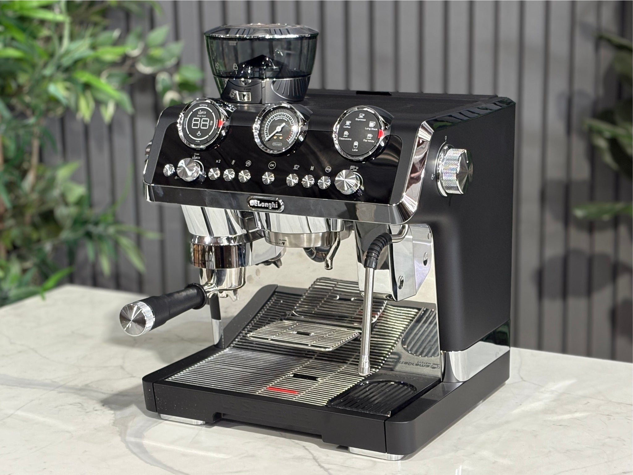 A DeLonghi La Specialista Maestro 1 Group Coffee Machine in Black is sitting on a marble bench. There is greenery in the background. The image is taken from the front of the machine and the machine is sitting on an angle so that you can see the front of the machine and the side. 