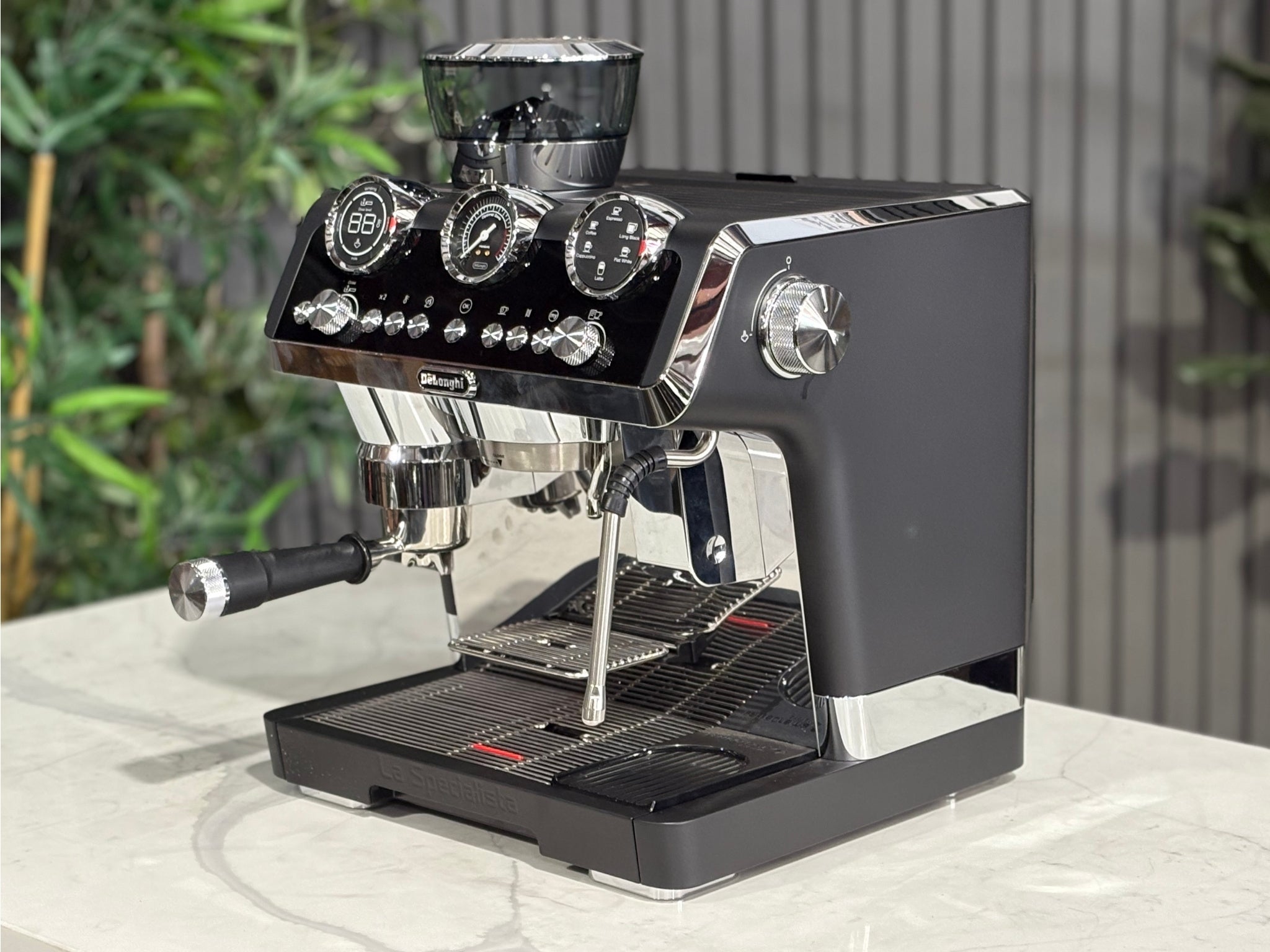 A DeLonghi La Specialista Maestro 1 Group Coffee Machine in Black is sitting on a marble bench. There is greenery in the background. The image is taken from the front of the machine and the machine is sitting on an angle so that you can see the front of the machine and the side. 