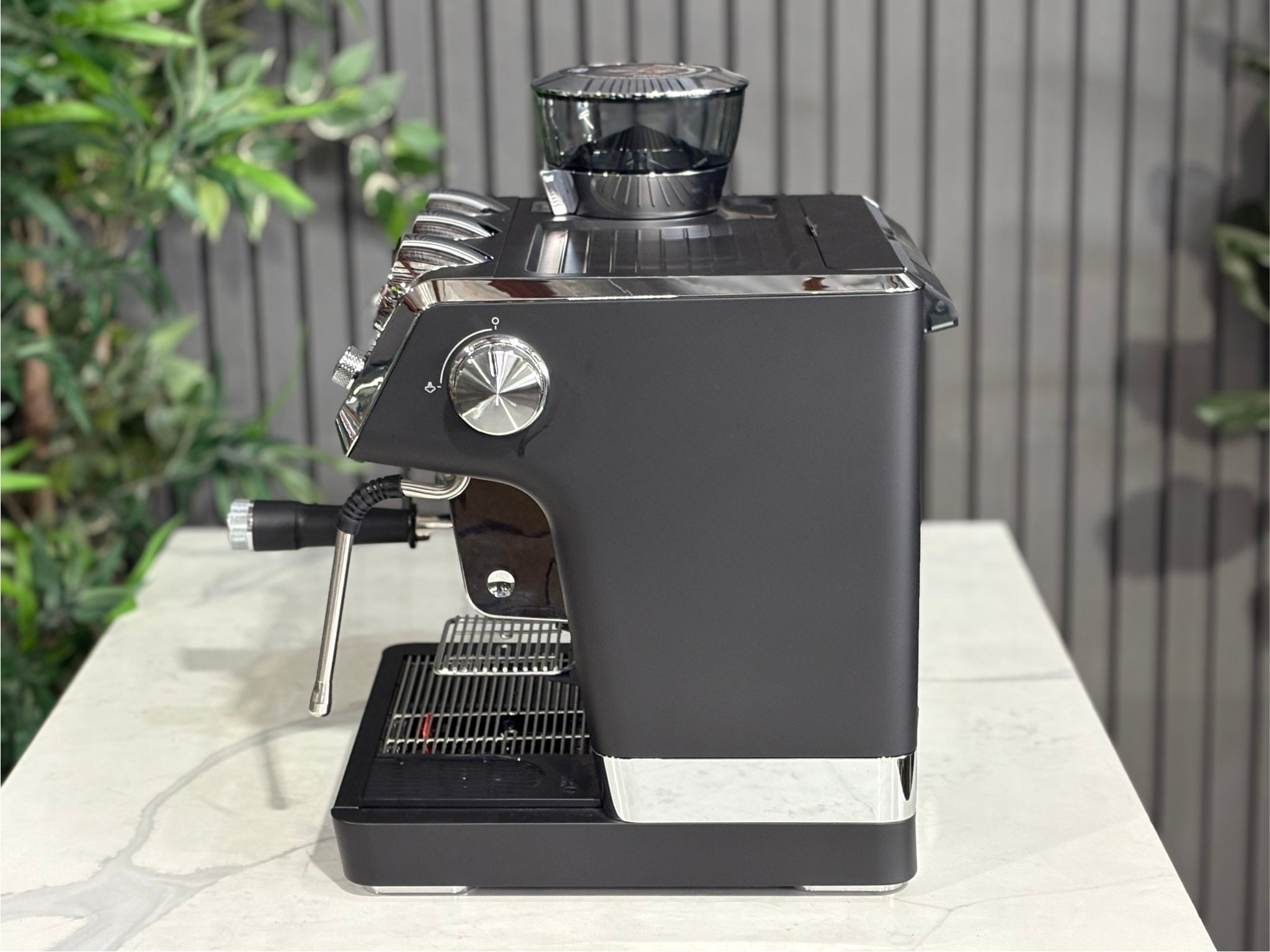 A DeLonghi La Specialista Maestro 1 Group Coffee Machine in Black  sitting on a marble bench. There is greenery in the background. The image is taken from the side of the machine.
