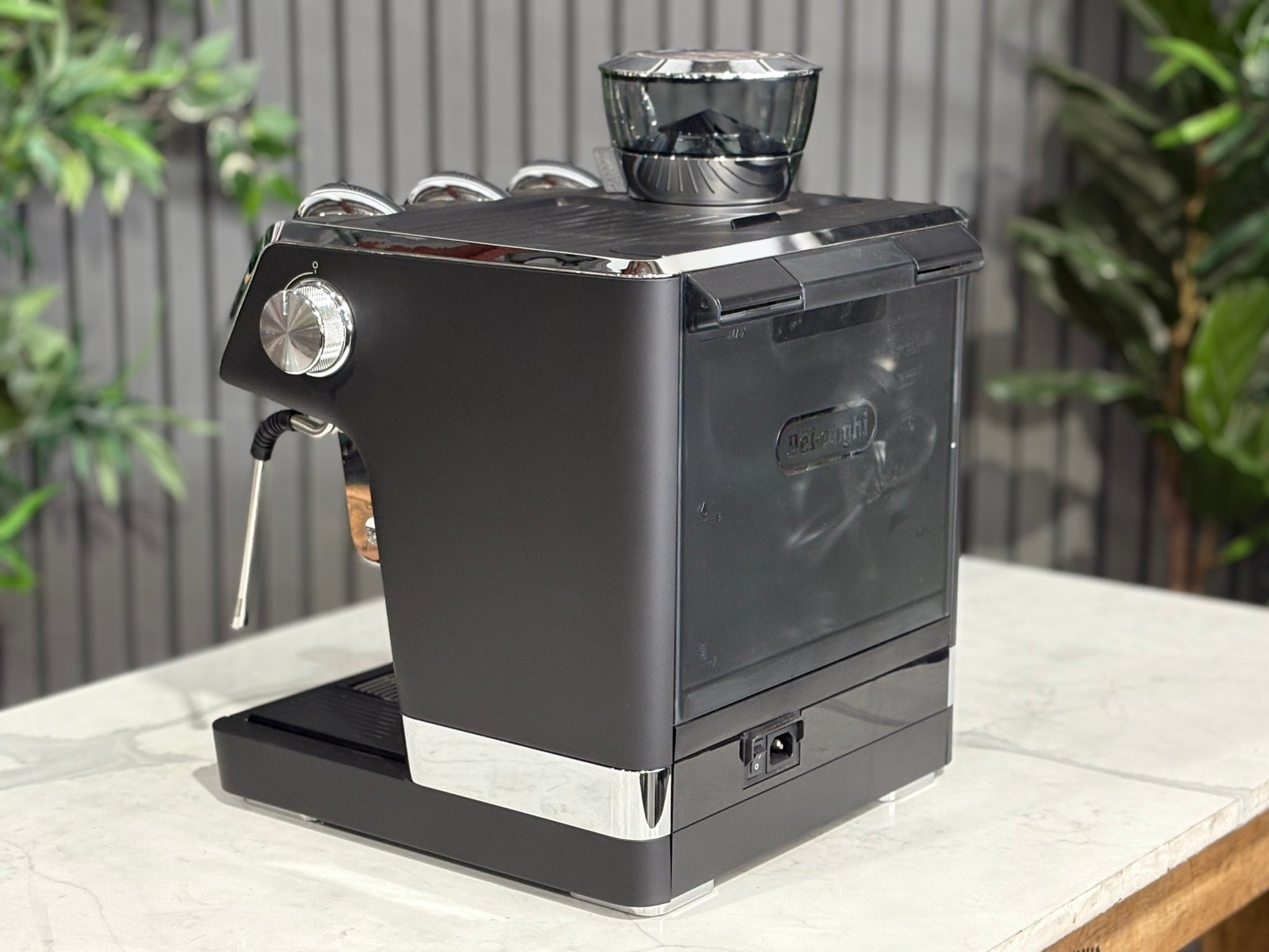 A DeLonghi La Specialista Maestro 1 Group Coffee Machine in Black sitting on a marble bench. There is greenery in the background. The image is taken from behind the machine and the machine is sitting on an angle so that you can see the back of the machine and the side.