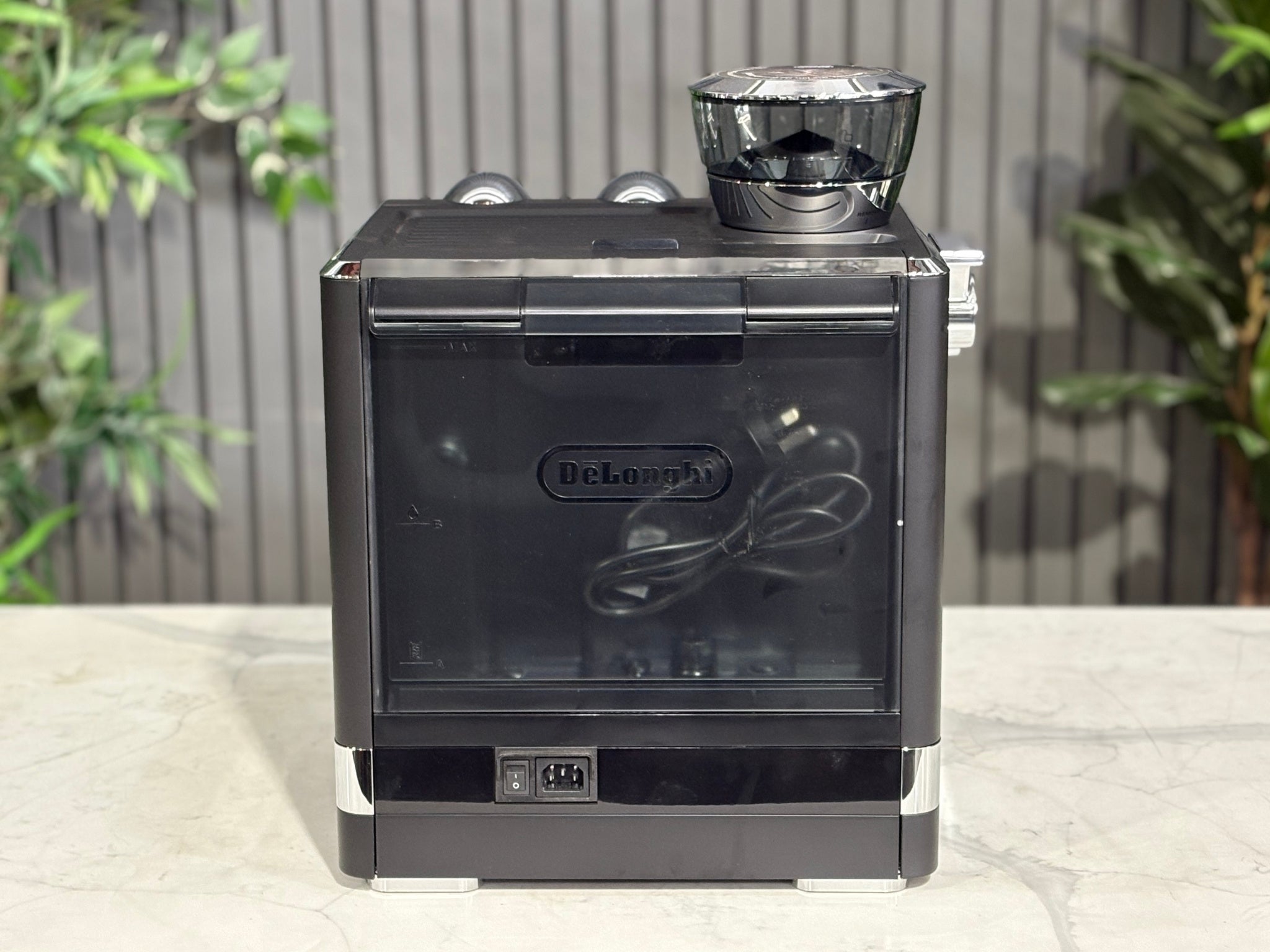 A DeLonghi La Specialista Maestro 1 Group Coffee Machine in Black is sitting on a marble bench. There is greenery in the background. The image is taken from behind the machine so the back is visible