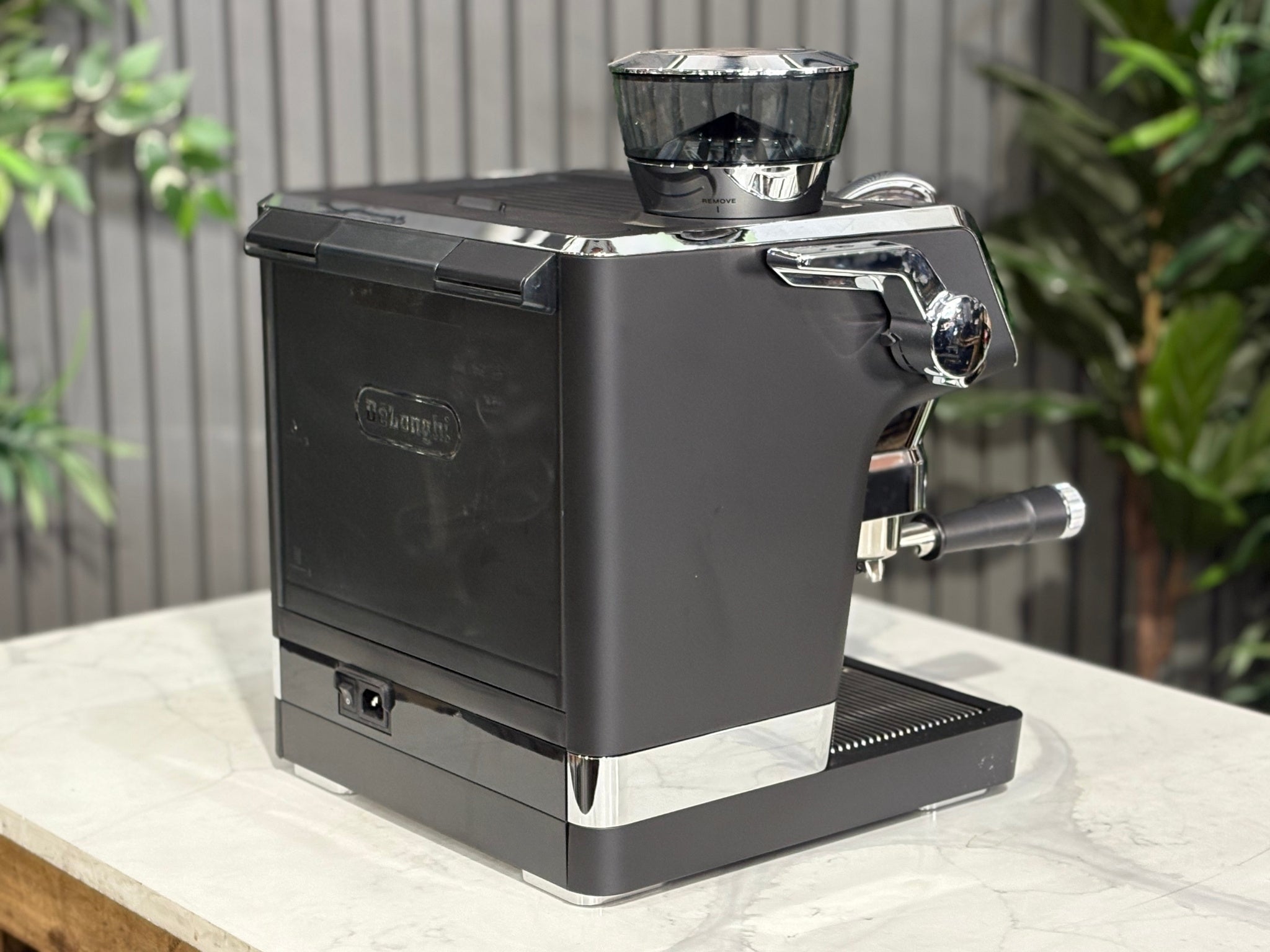 A DeLonghi La Specialista Maestro 1 Group Coffee Machine in Black sitting on a marble bench. There is greenery in the background. The image is taken from behind the machine and the machine is sitting on an angle so that you can see the back of the machine and the side.