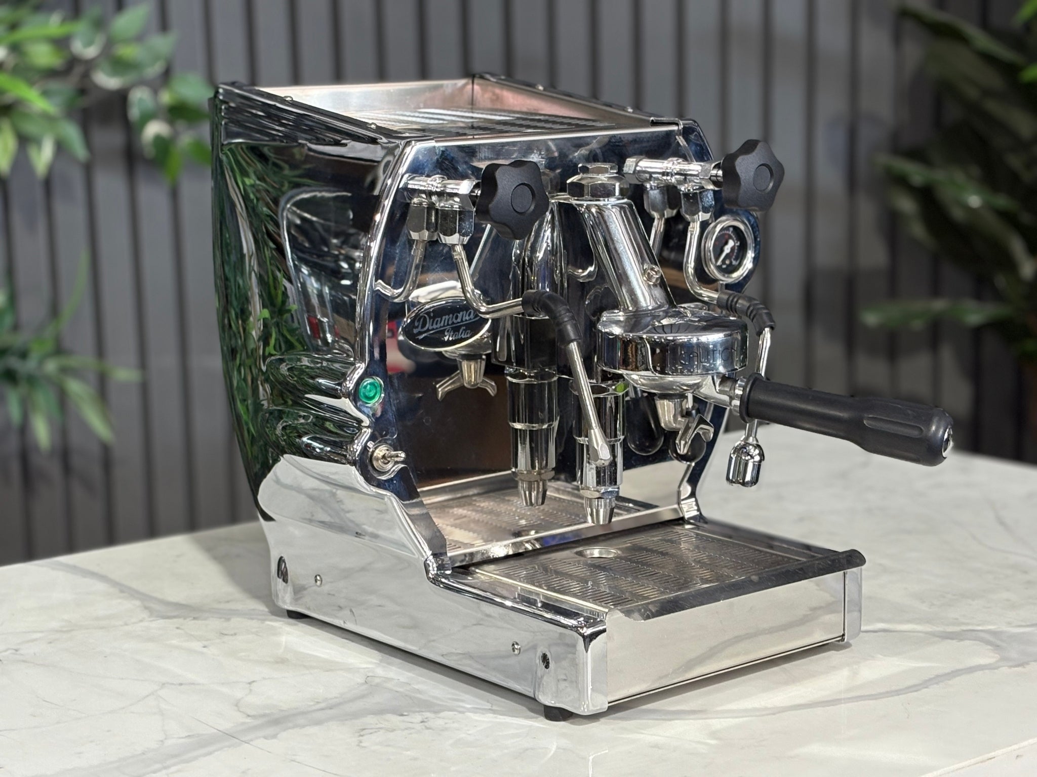 A Diamond Cuadra 1 Group Espresso Coffee Machine with Stainless finish is sitting on a marble bench. There is greenery in the background. The image is taken from the front of the machine and the machine is sitting on an angle so that you can see the front of the machine and the side. 