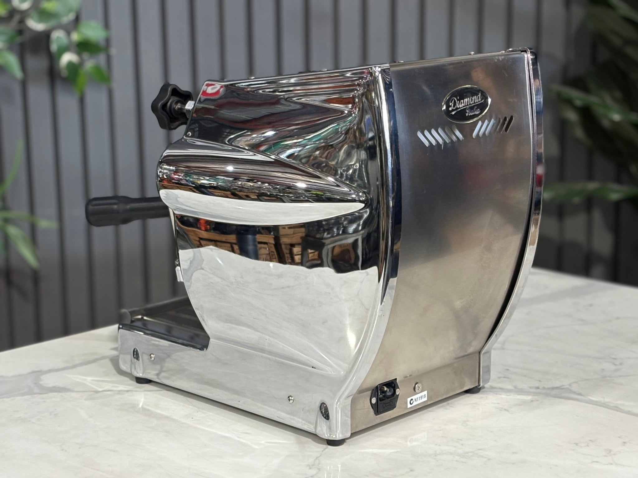 A Diamond Cuadra 1 Group Espresso Coffee Machine with Stainless finish itting on a marble bench. There is greenery in the background. The image is taken from behind the machine and the machine is sitting on an angle so that you can see the back of the machine and the side.